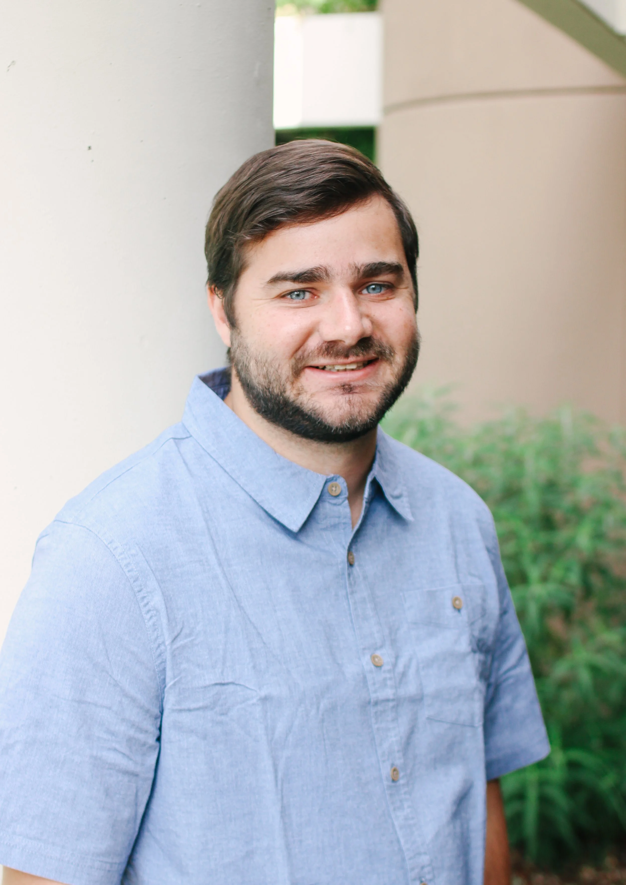 Portrait of a man with dark hair and a beard, wearing a blue button-up shirt, outdoors with trees and a fence in the background.