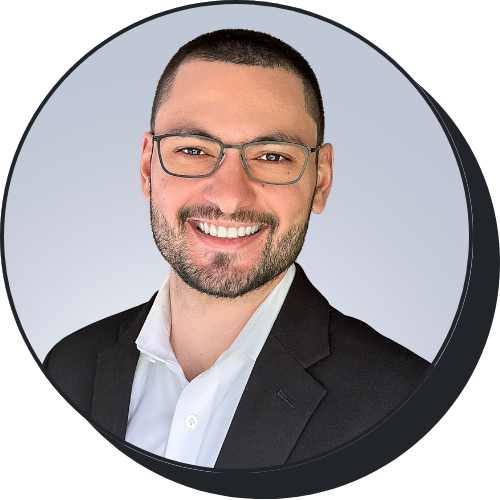 Headshot of a smiling man with short dark hair, glasses, a beard, wearing a black suit and white shirt, against a light gray background.