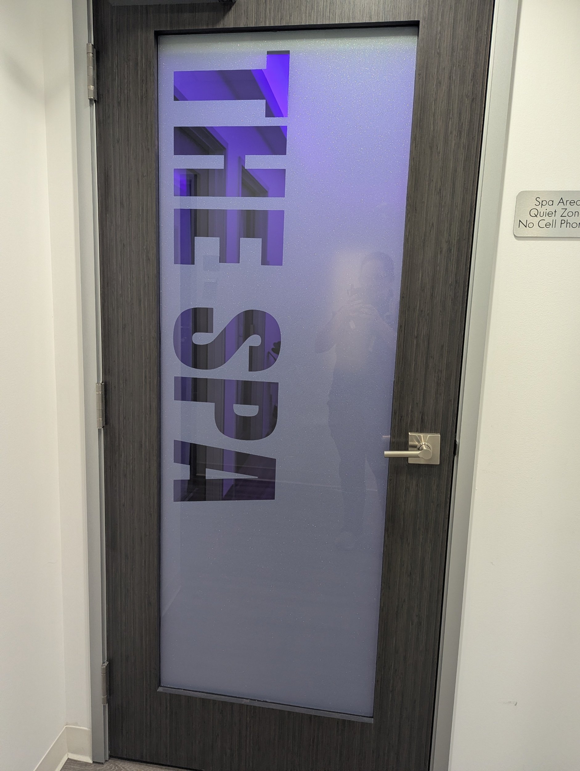 A door with a frosted glass panel, featuring the word 'spa' in large letters and a purple LED strip lighting inside, located in a corridor with light-colored walls and a sign on the wall indicating a spa area, quiet zone, and no cell phones allowed.