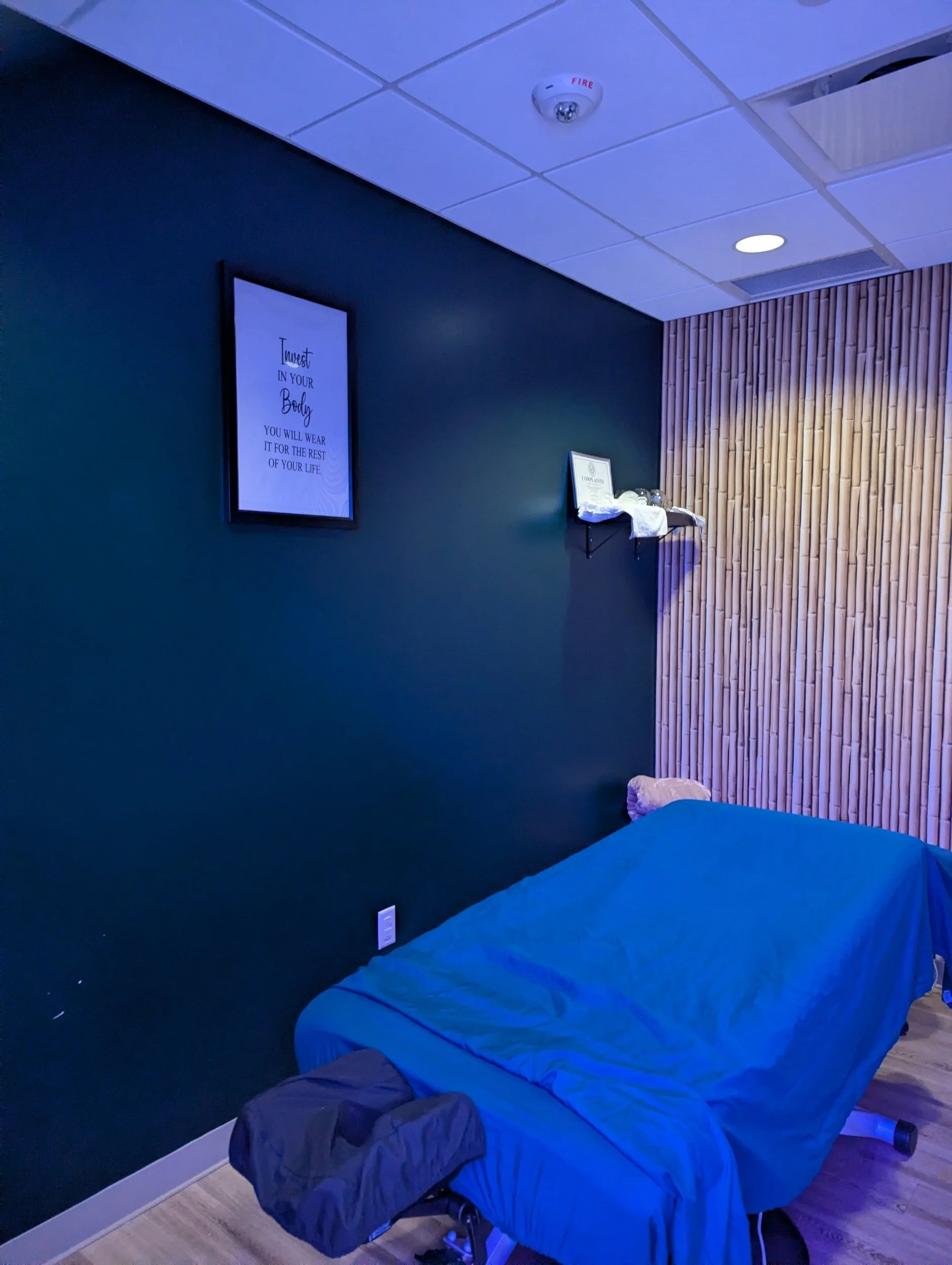 A massage or therapy room with a massage table covered in a blue sheet, dark green wall with framed quote, and unique bamboo wall decor. The ceiling has a fire alarm and overhead lighting.