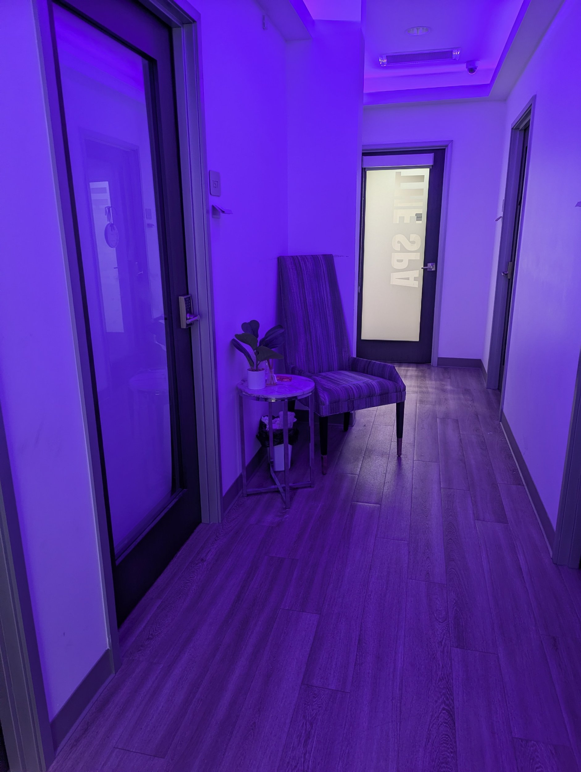 An indoor hallway illuminated with purple lighting, featuring a massage room on the left, a chair with a striped pattern and a small side table with a plant on it, and a door with frosted glass that has the words 'Spa Area' written on it.