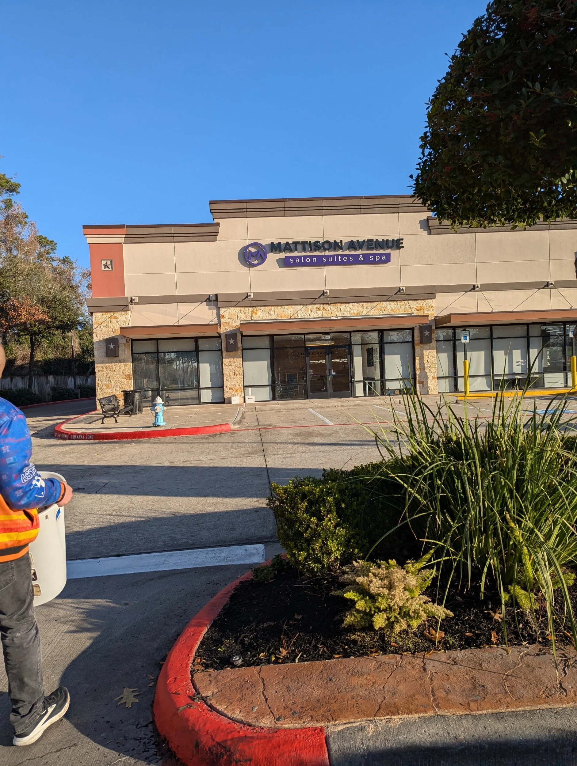 The exterior of a Salon Suites and Spa building called Mattison Avenue, with a sidewalk, a small landscaped area, and a person standing in the foreground. The building has beige walls and a sign with purple and black lettering.