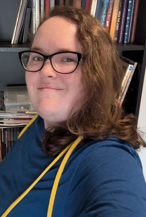 A person with long curly hair and glasses smiling, wearing a blue shirt with yellow resistance band, standing in front of a bookshelf filled with books.