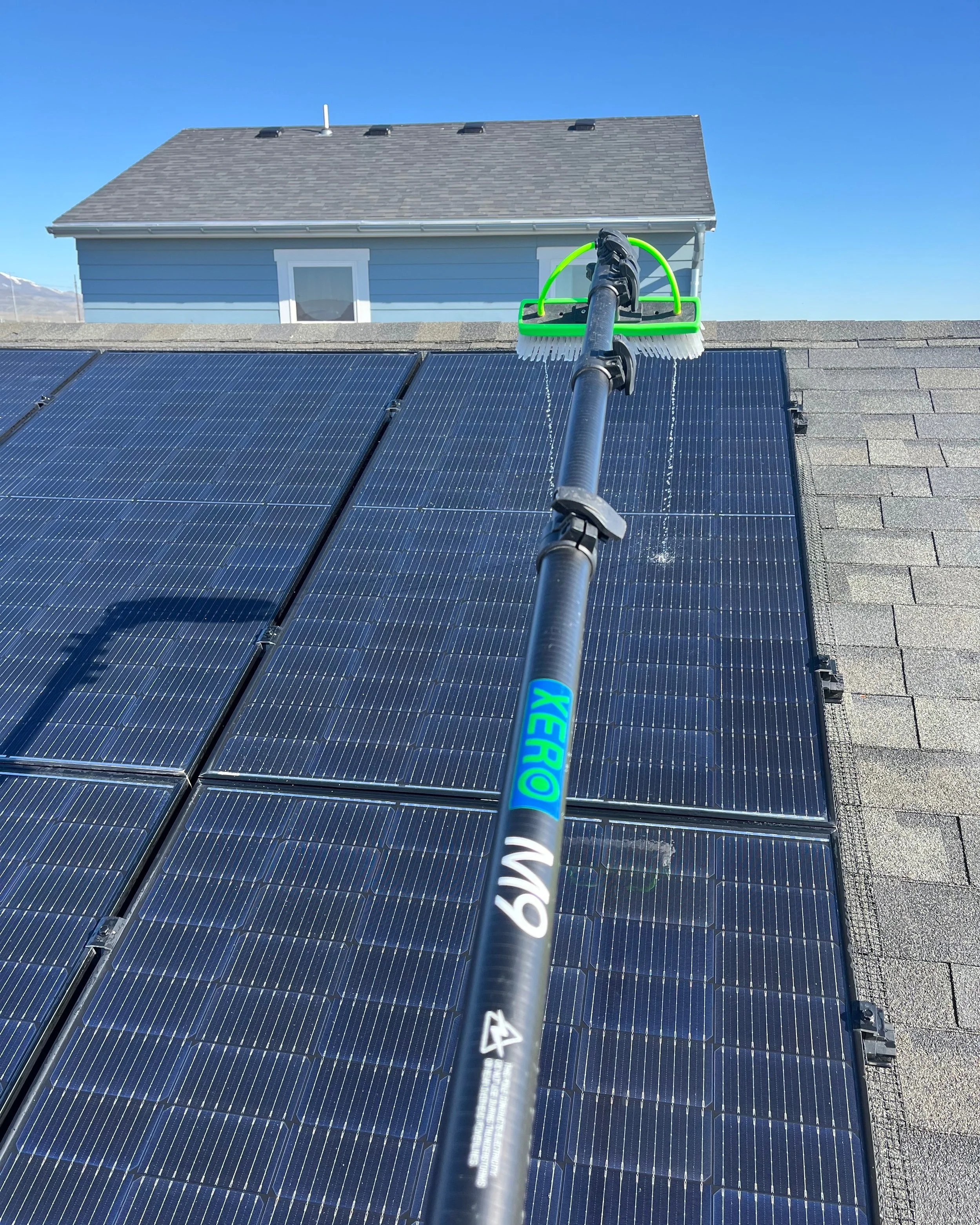 View from a roof showing solar panels being cleaned with a long-handled brush, with a house and clear blue sky in the background.