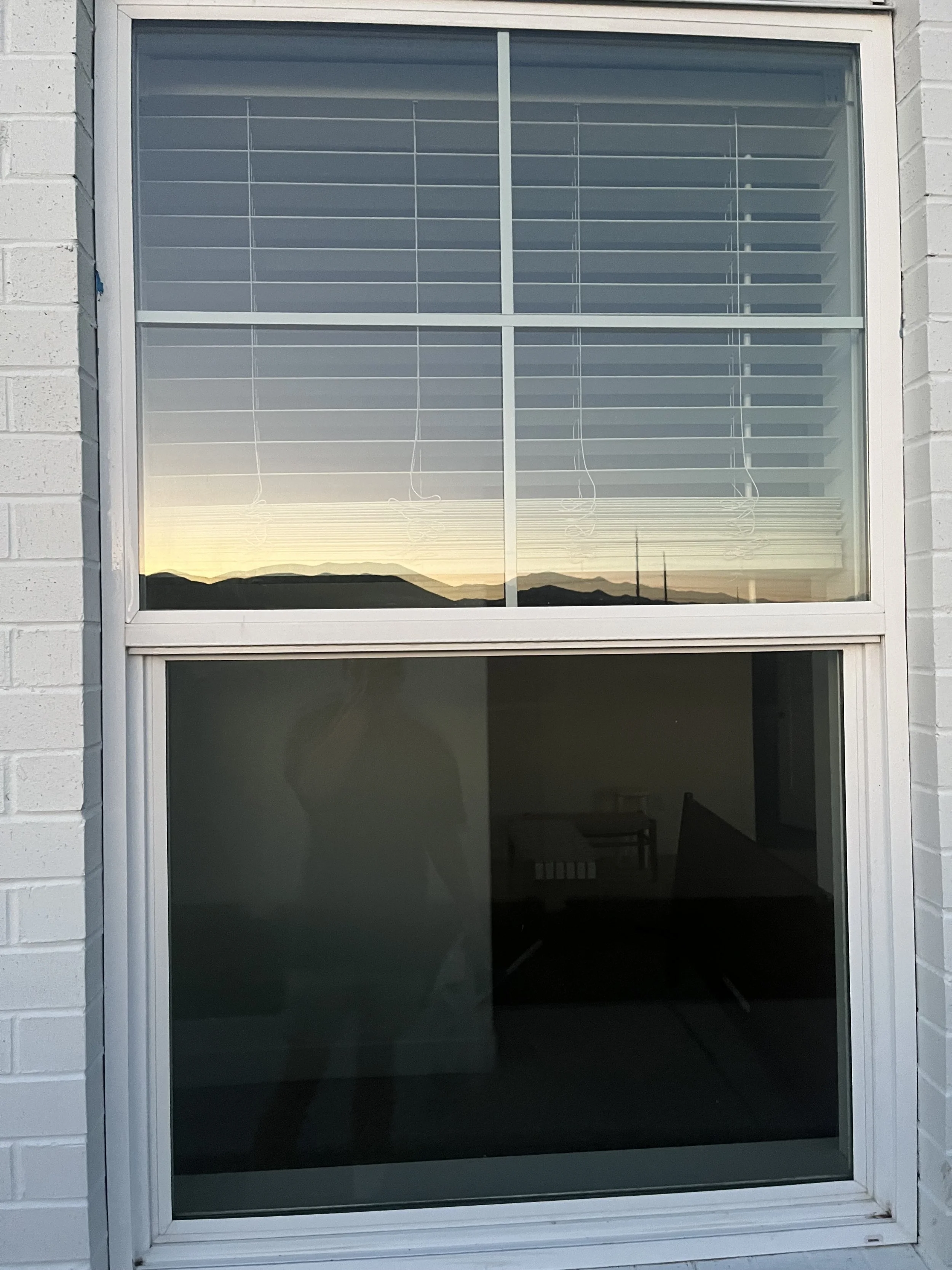 A double-pane window with white blinds on the upper window and a reflection of mountains and the sunset, while the lower window shows a darker interior with furniture.