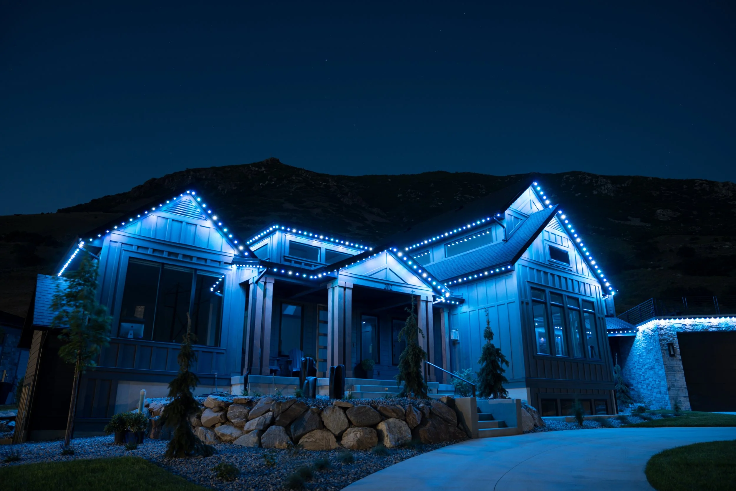 Night view of a modern house with blue exterior lighting and a landscaped yard with rocks and small trees.