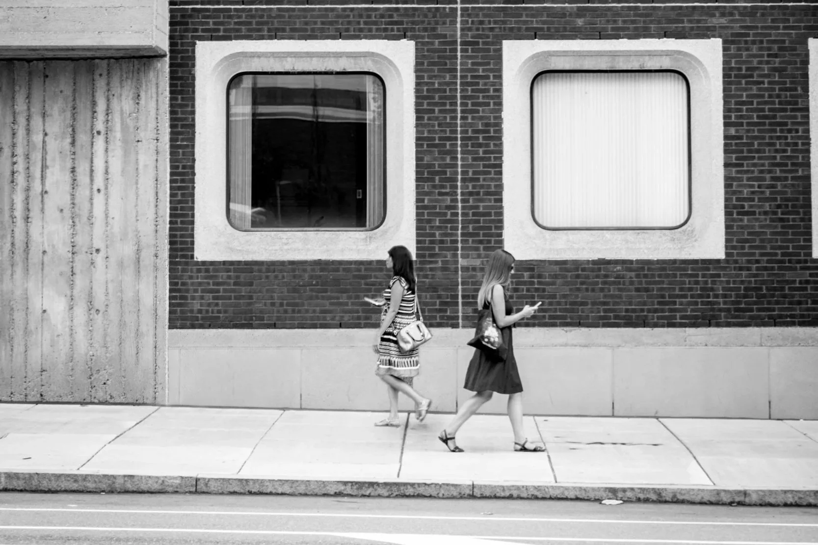 Two women walking on a sidewalk in front of a brick wall with two large windows, all in black and white.