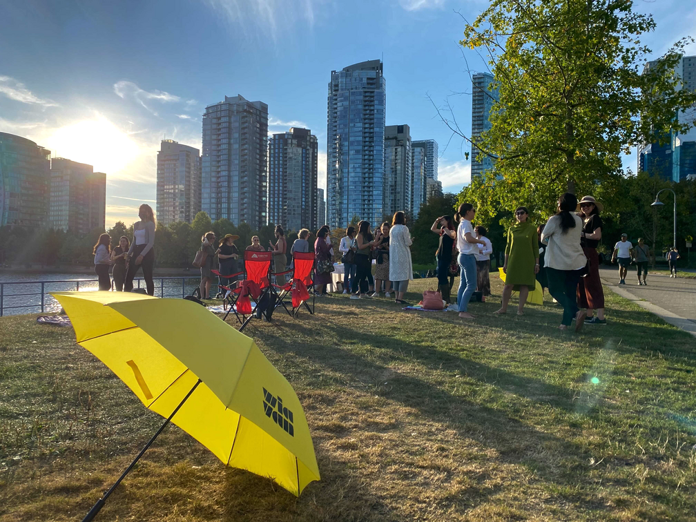 A group of people gathered outdoors near a waterway with high-rise buildings in the background. The sun is setting, casting long shadows. A yellow umbrella is on the ground in the foreground.