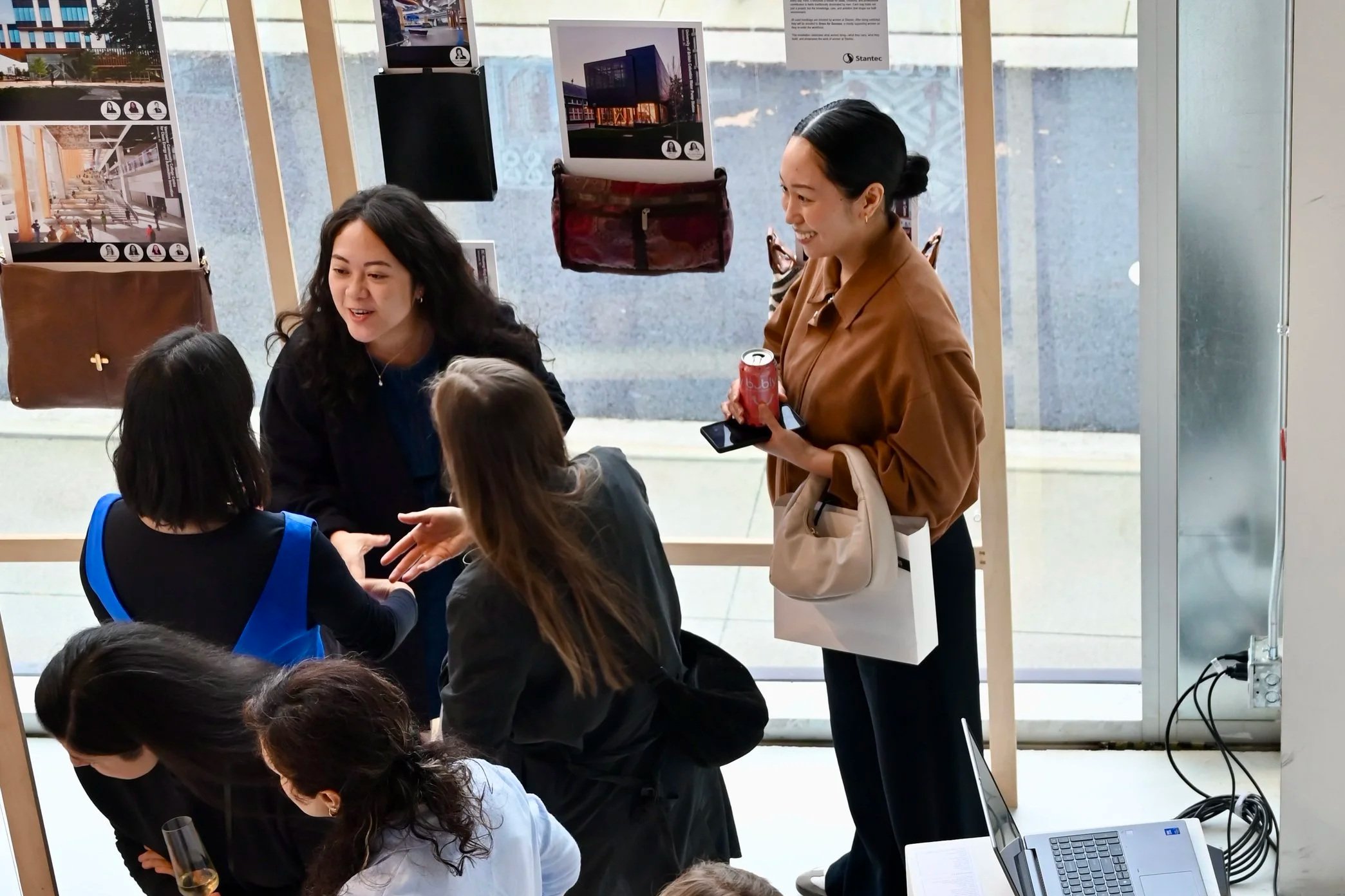 A group of women and girls gathered at an event or exhibition, talking and holding drinks. One woman is holding a phone and a soda can, smiling. The background shows display panels with architectural renderings, handbags hanging on a stand, and a laptop on a table.