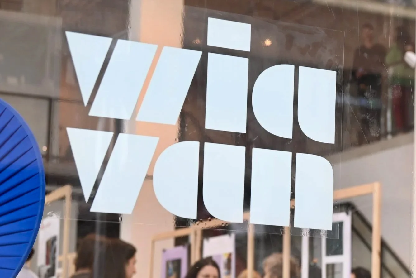 Sign with the word "Vitra" written in stylized white letters on a transparent background, reflecting the interior of a shopping mall or store.