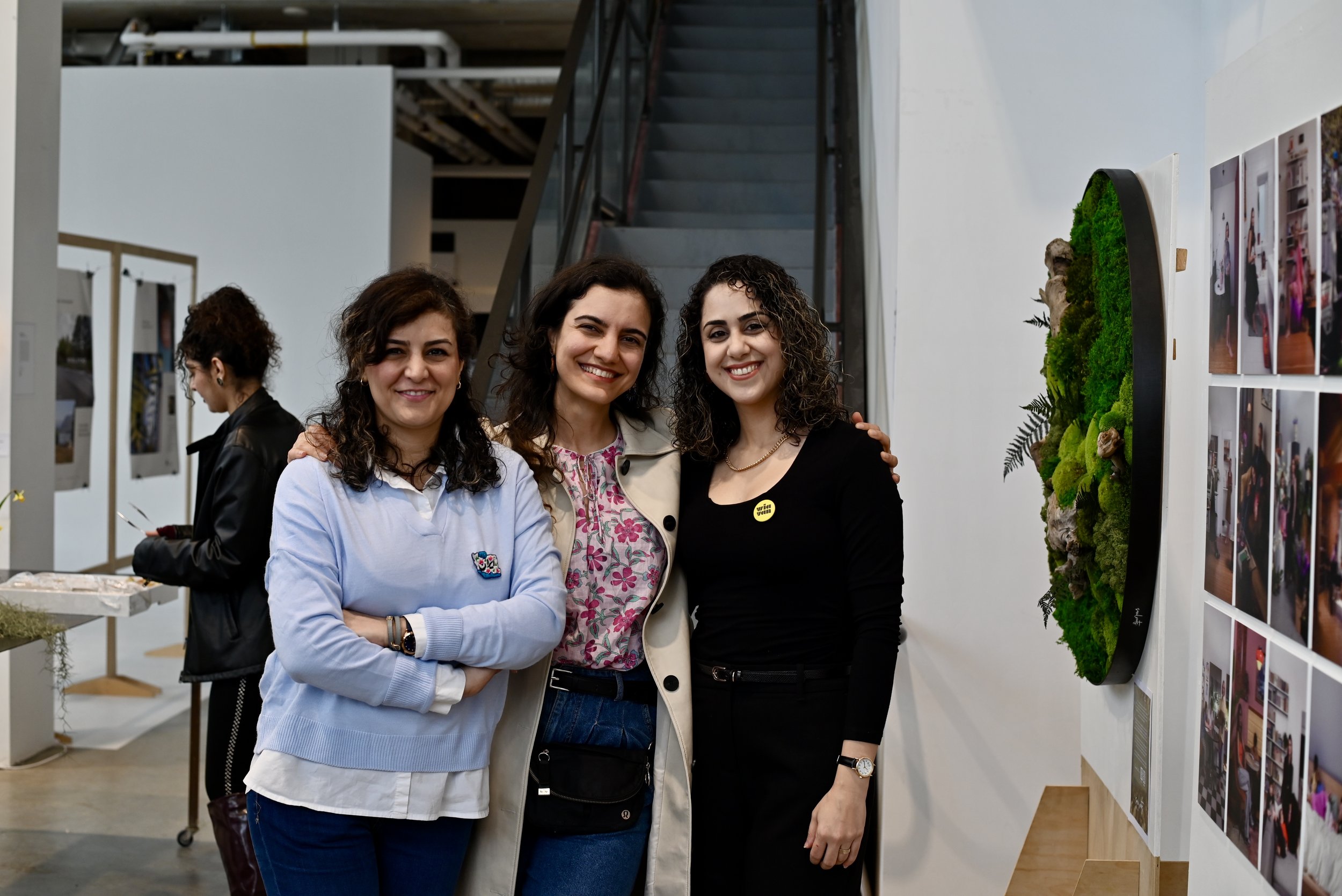 Three women standing close together at an indoor exhibition, smiling at the camera. There are photos on the wall to the right and display tables in the background.