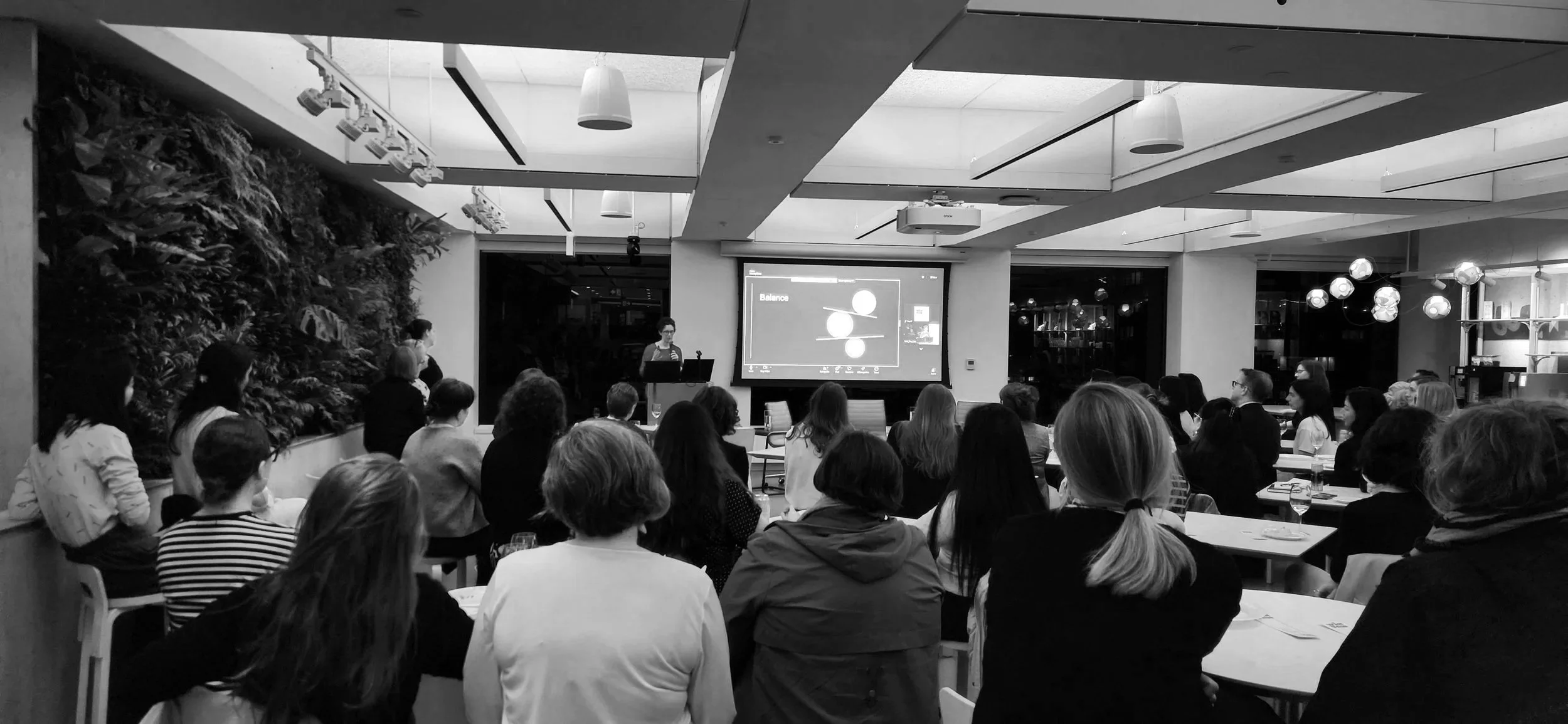 A group of people attending a presentation in a modern conference room with a speaker at a podium and a large screen displaying a diagram.