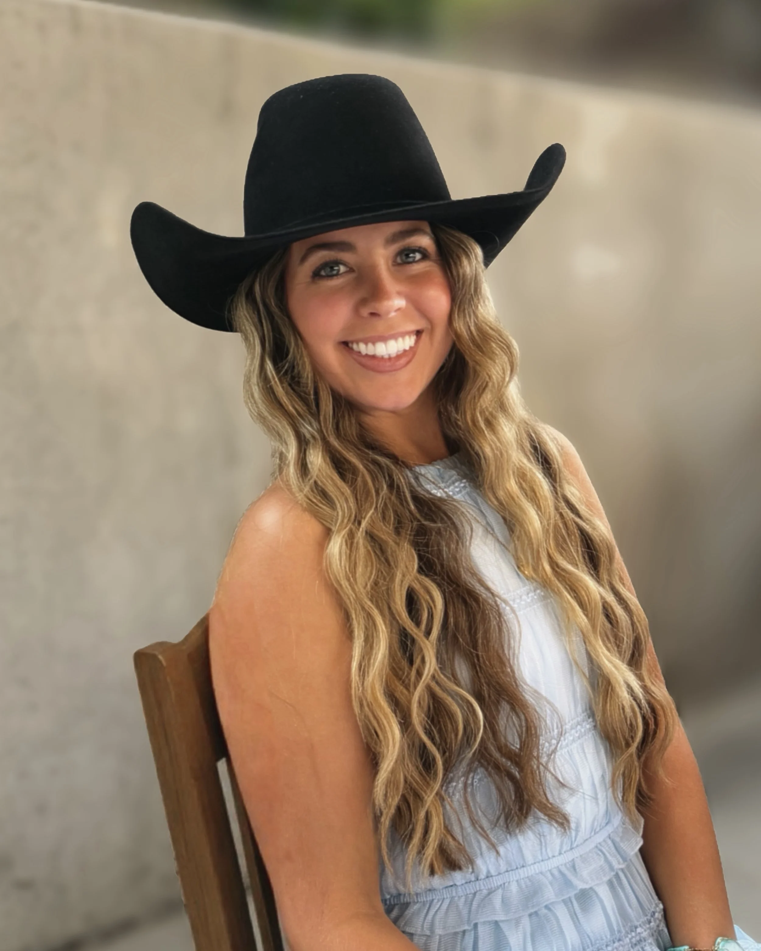 A smiling woman with long curly hair wearing a wide-brimmed black hat and a light-colored sleeveless top sits on a wooden chair.