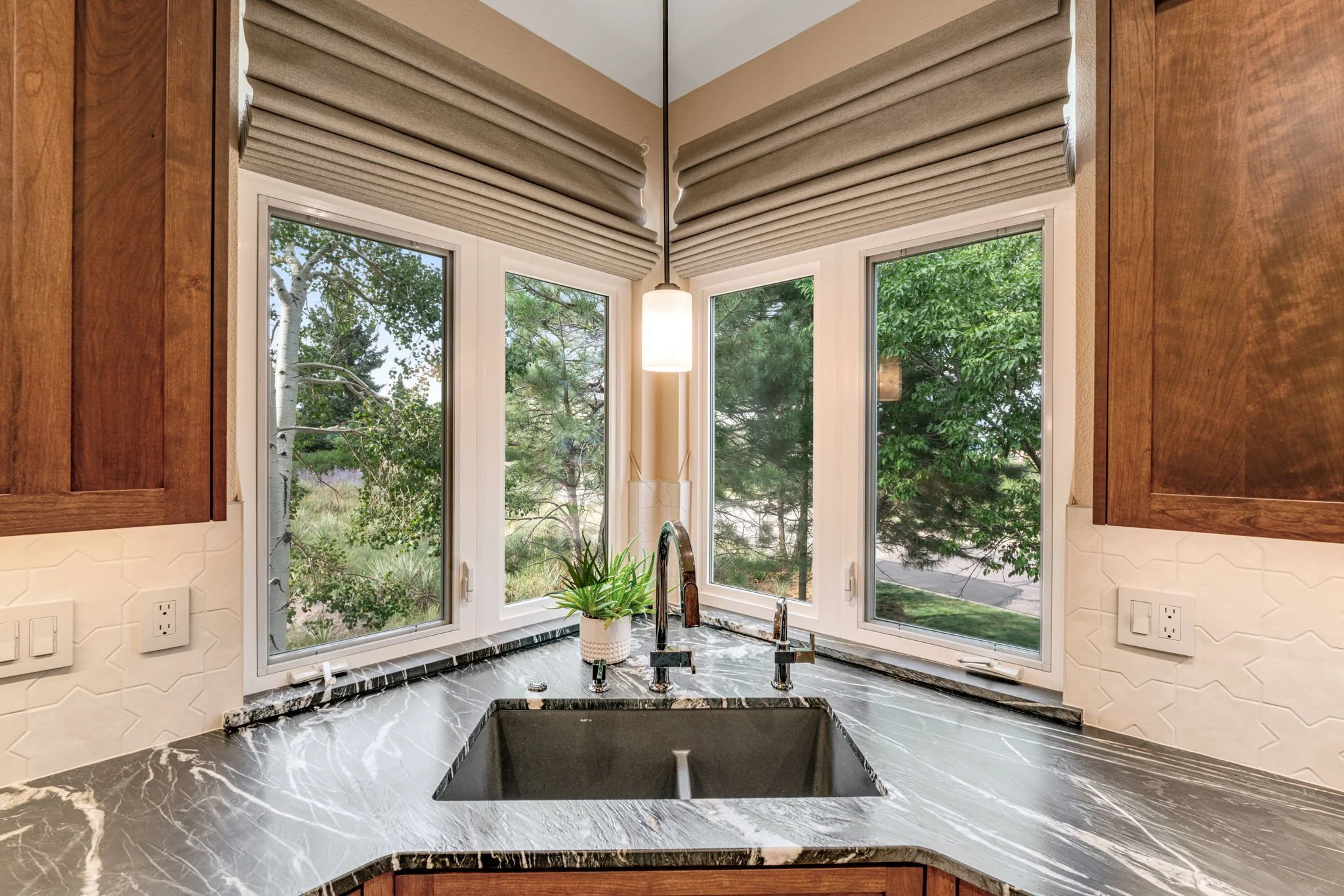 Kitchen window area with three large windows showing green trees outside, beige Roman shades partially drawn, a black marble countertop with a sink and faucet, a potted plant, and wood cabinets on each side.