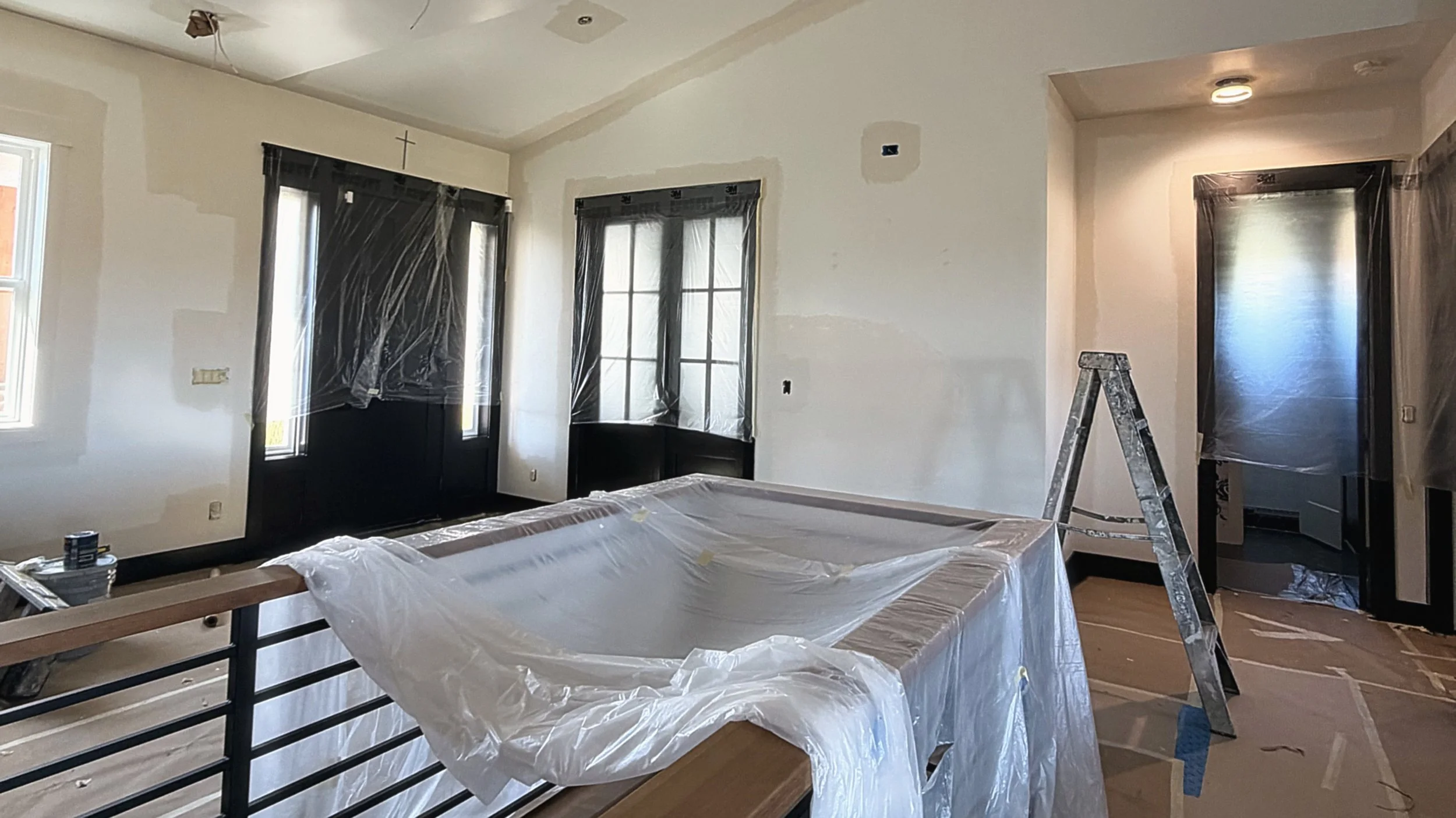 Interior room under renovation with plastic-covered furniture, black window and door frames, a painted wall, ladder, and construction materials.