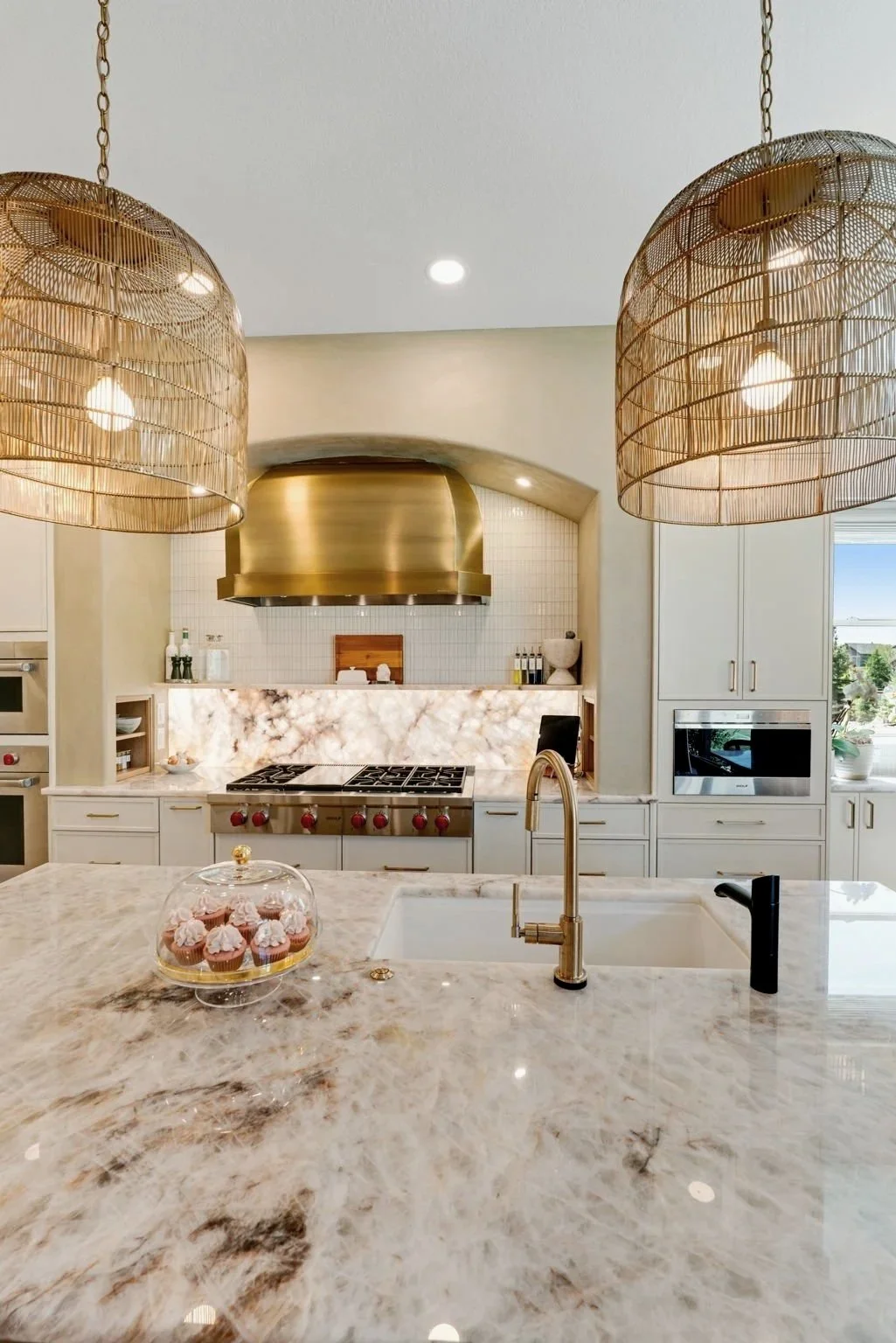 Modern kitchen with a marble island, gold range hood, white cabinetry, and large woven pendant lights.