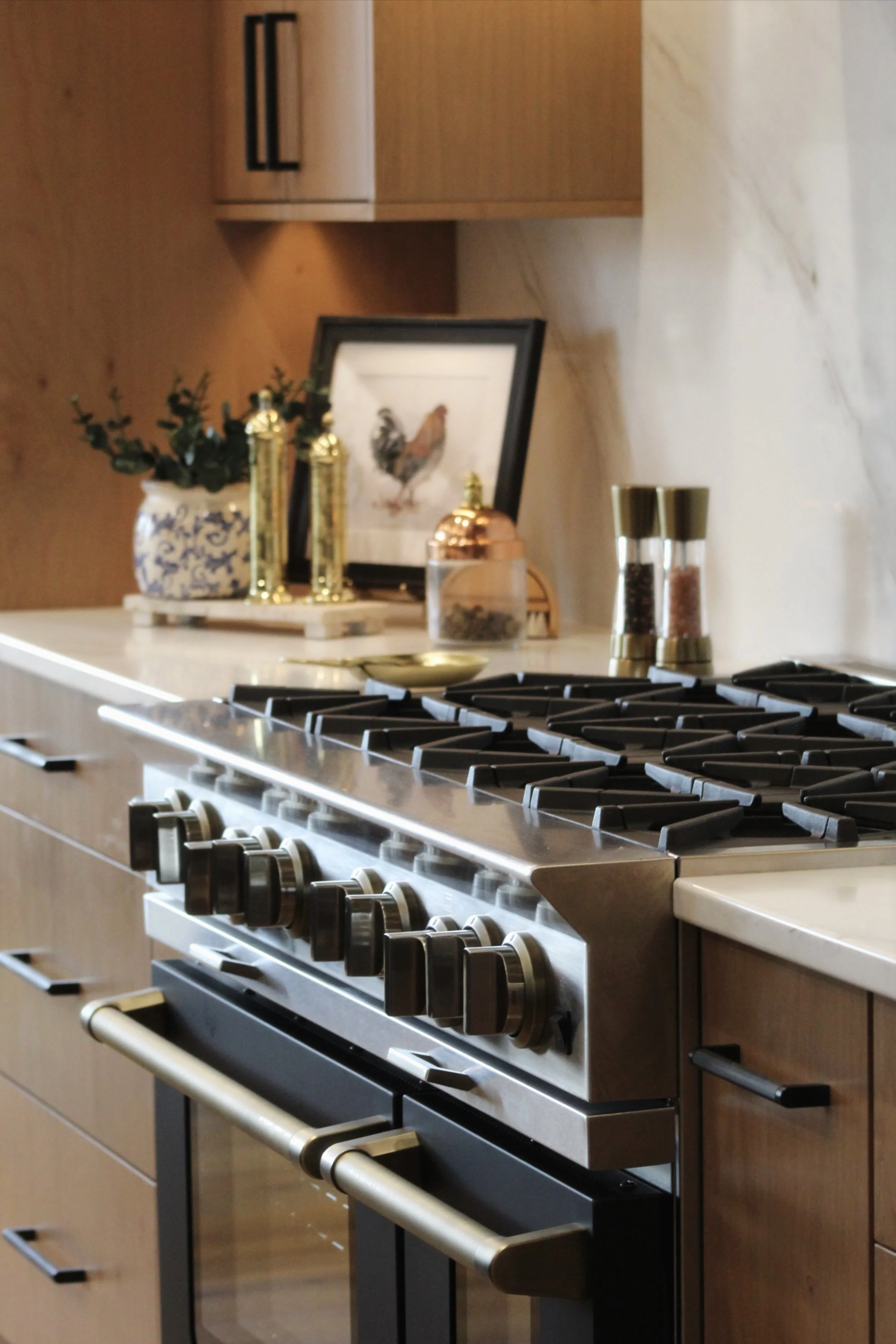 Stainless steel gas stove with black burners and control knobs, set in a wooden kitchen with a white marble backsplash. Decorative items on the countertop include a framed picture of a rooster, a white and blue vase with greenery, two tall gold-colored containers, and spice shakers.