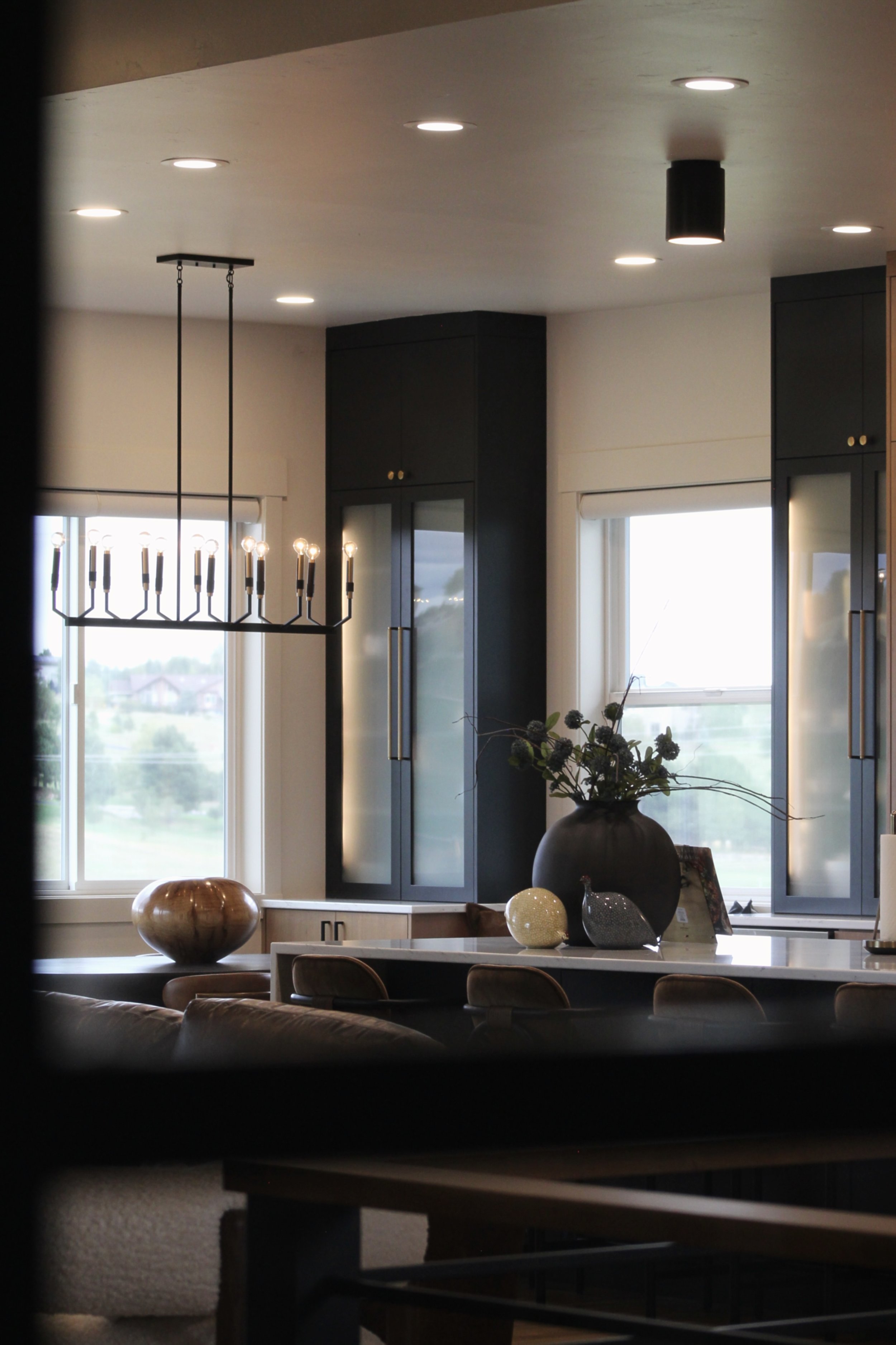 A modern kitchen with dark blue cabinets, large windows, and a marble countertop. Decorative vases and a large potted plant are on the counter. A black chandelier with multiple candle-like lights hangs from the ceiling.