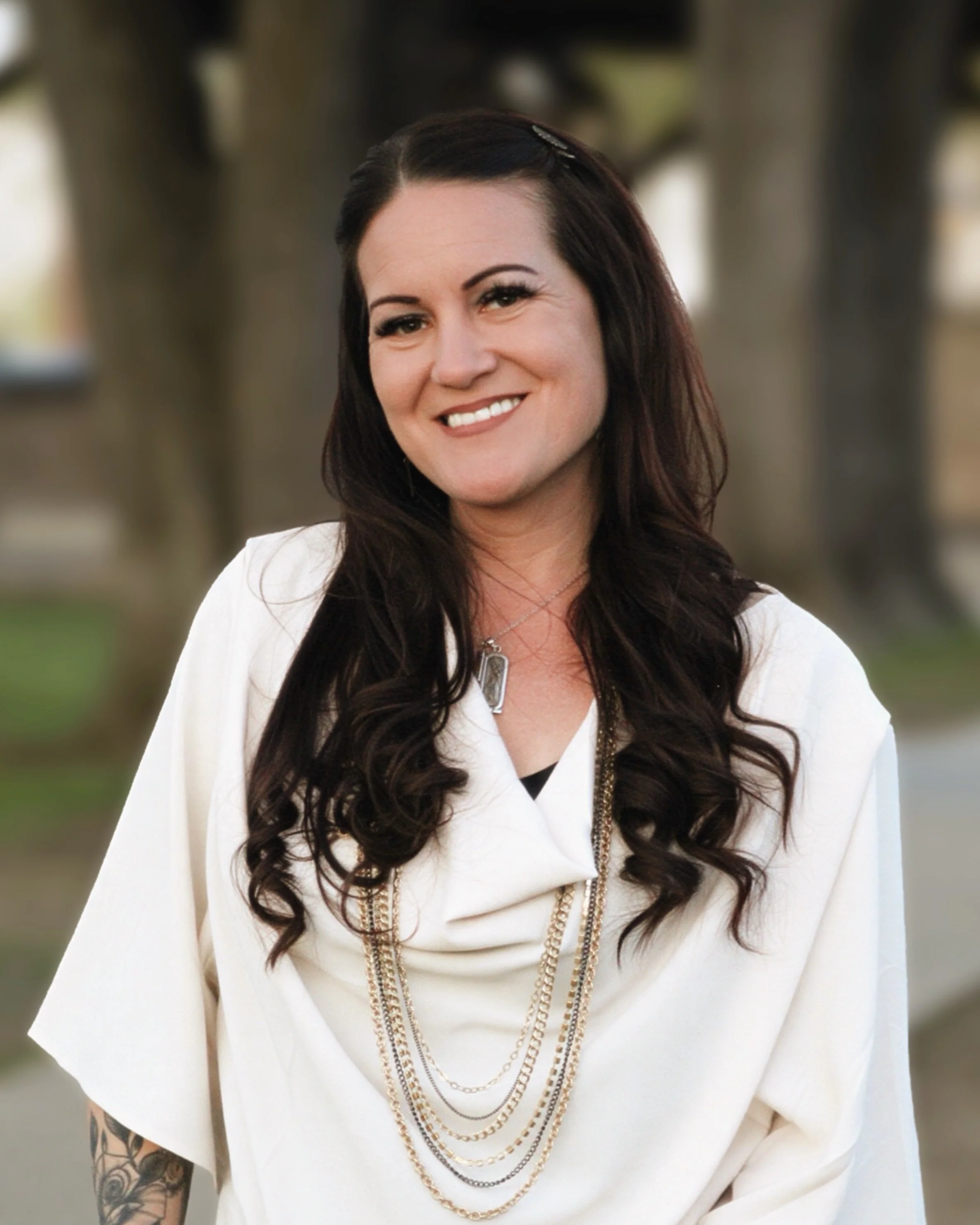 A woman with long dark hair, smiling, wearing a white shirt with layered gold chains, standing outdoors with blurred trees in the background.