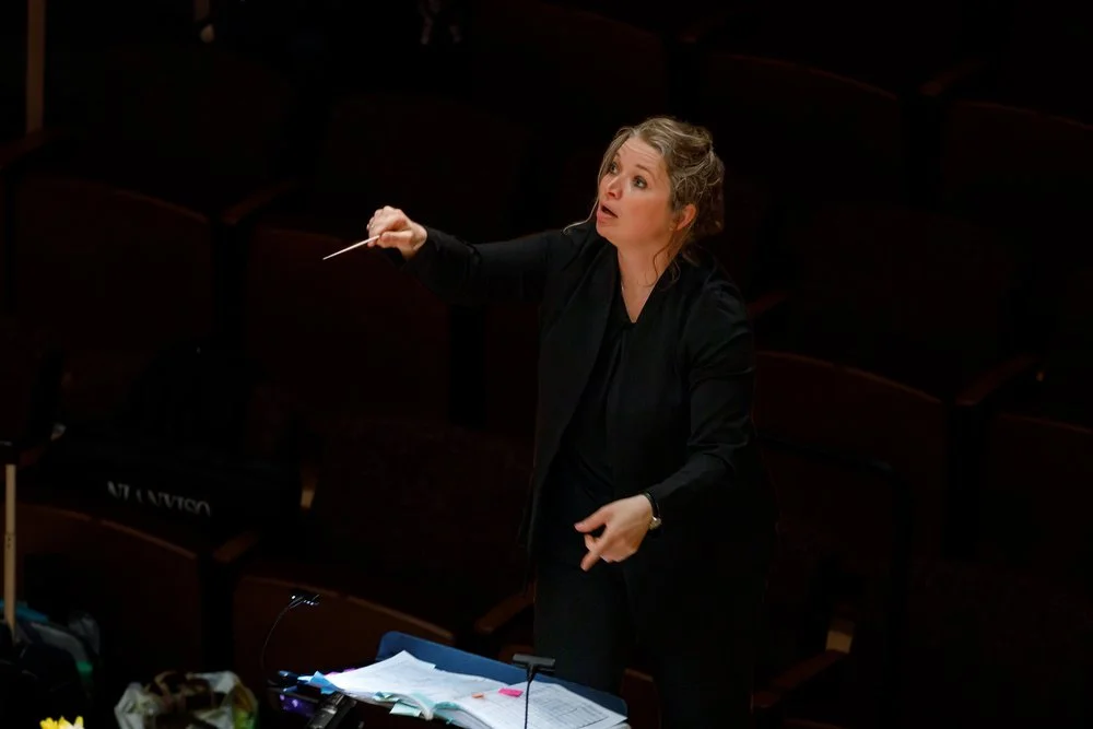 Orchestra conductor leading rehearsal in an auditorium with rows of empty seats.