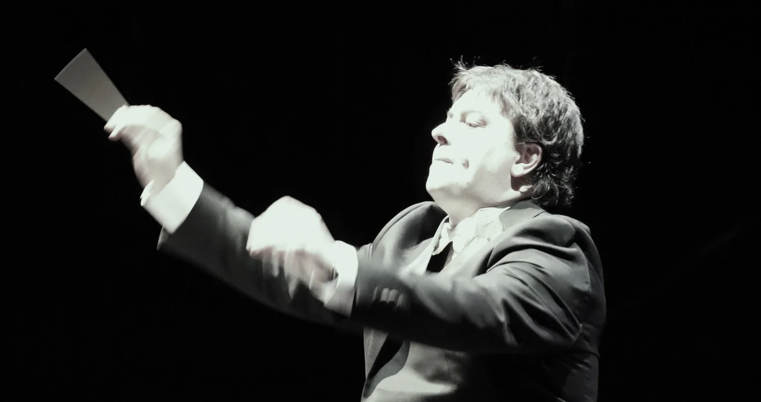 A male conductor in a suit leading an orchestra, with a focused expression, against a dark background.