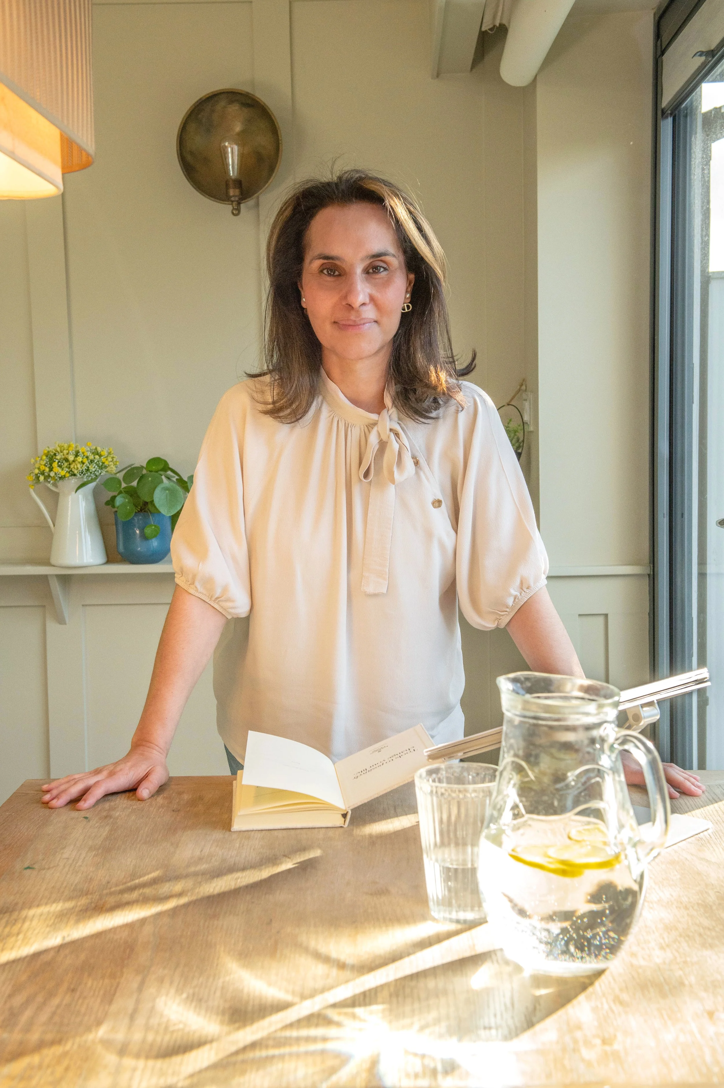 A woman with shoulder-length brown hair, wearing a cream-colored blouse with a bow, standing behind a wooden table with a glass pitcher of water, a glass, and an open book, in a sunlit room with potted plants and a window.