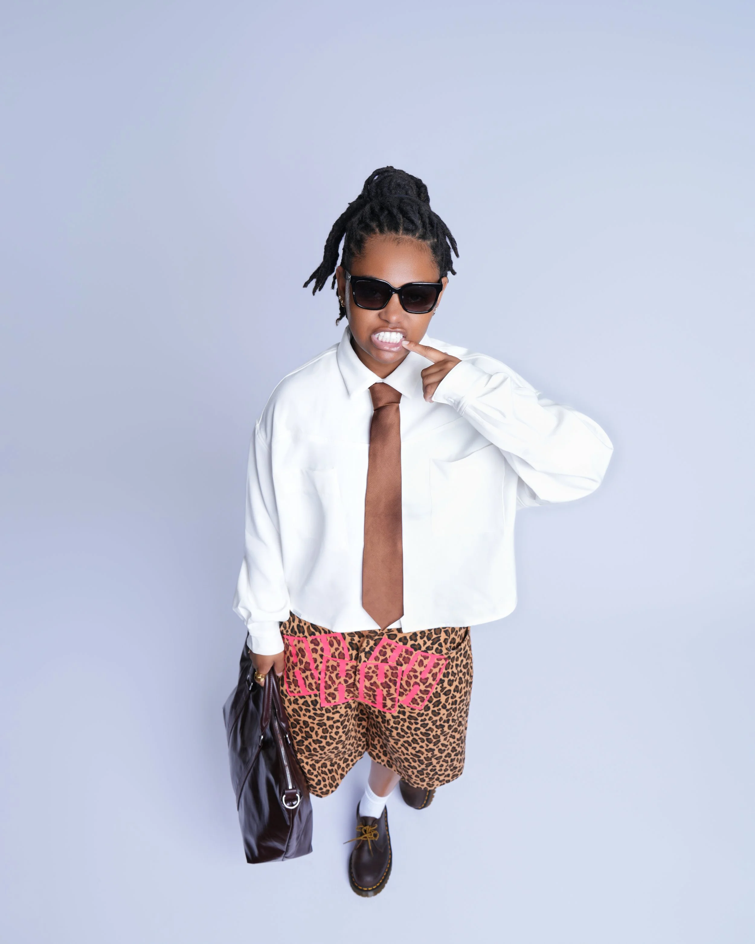 Person with dreadlocks wearing sunglasses, a white shirt, a brown tie, leopard print shorts with pink accents, standing against a light blue background.