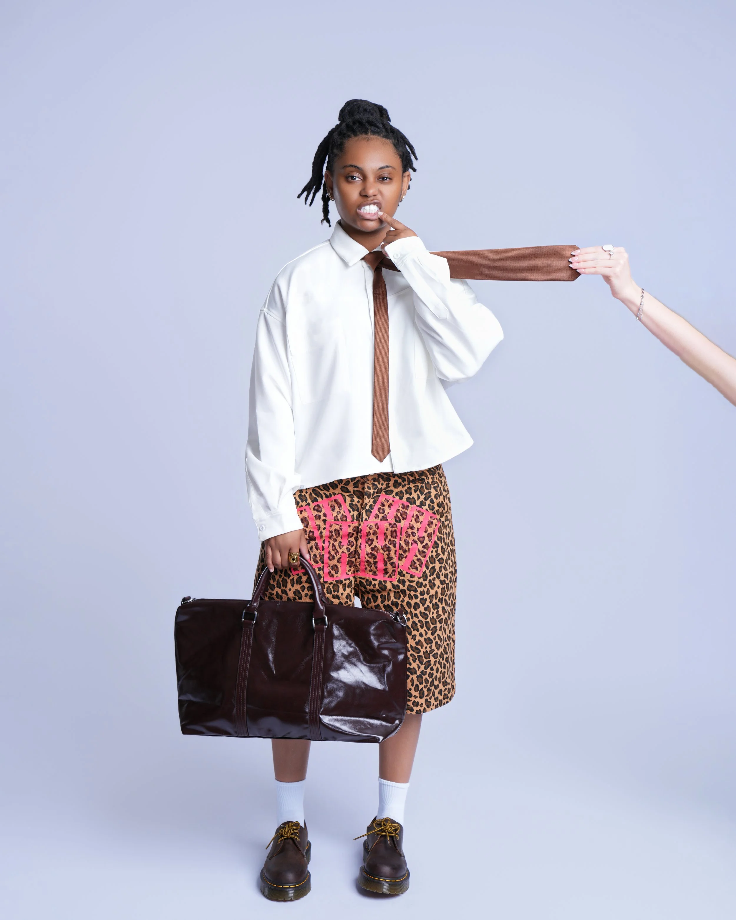 A woman with dreadlocks and a white shirt making a face while holding a large brown wooden stick against her shoulder, with a black bag in her hand and pink poker cards in front of her pants.