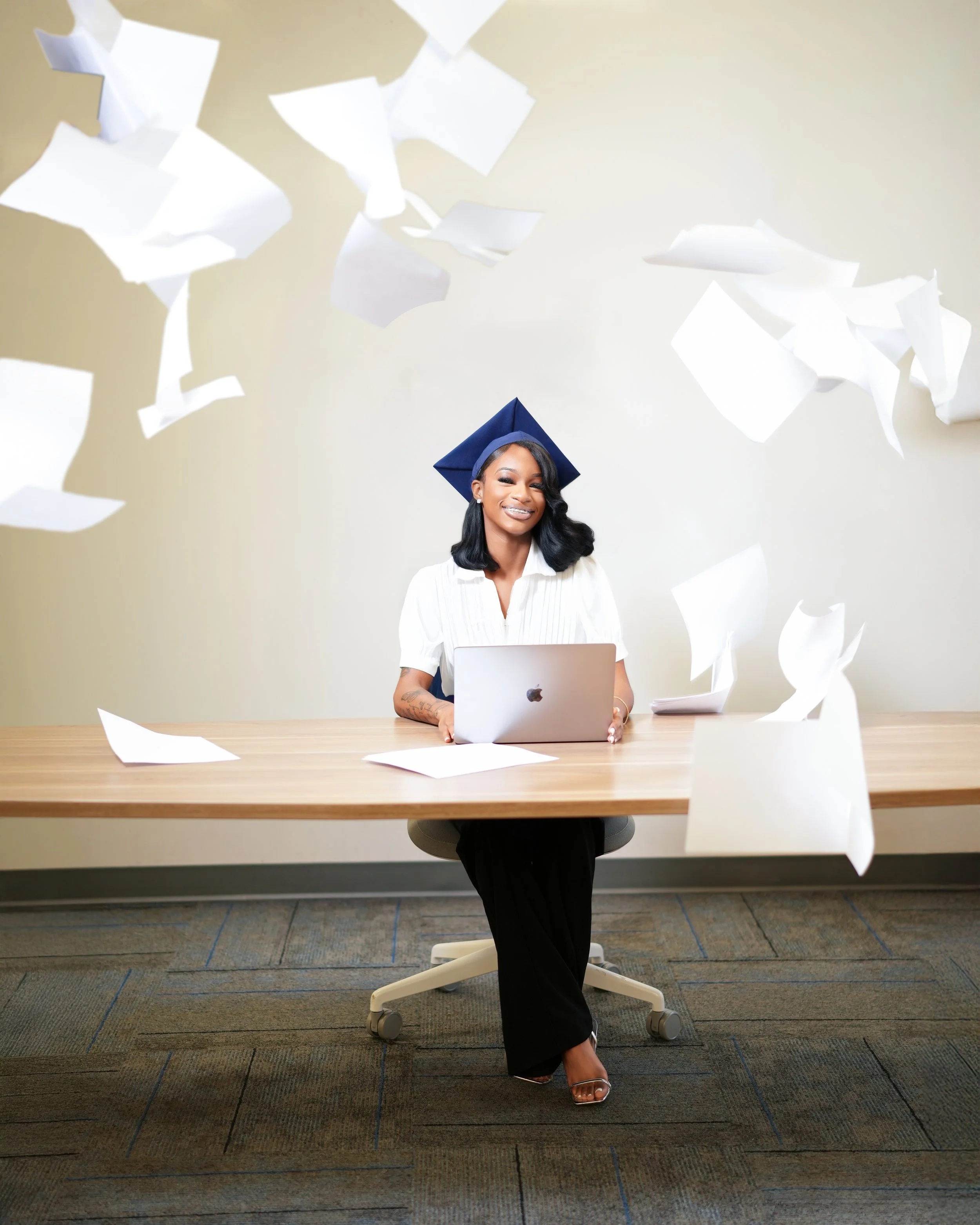 A woman in a graduation cap sits at a desk with a laptop, surrounded by flying sheets of paper, smiling.