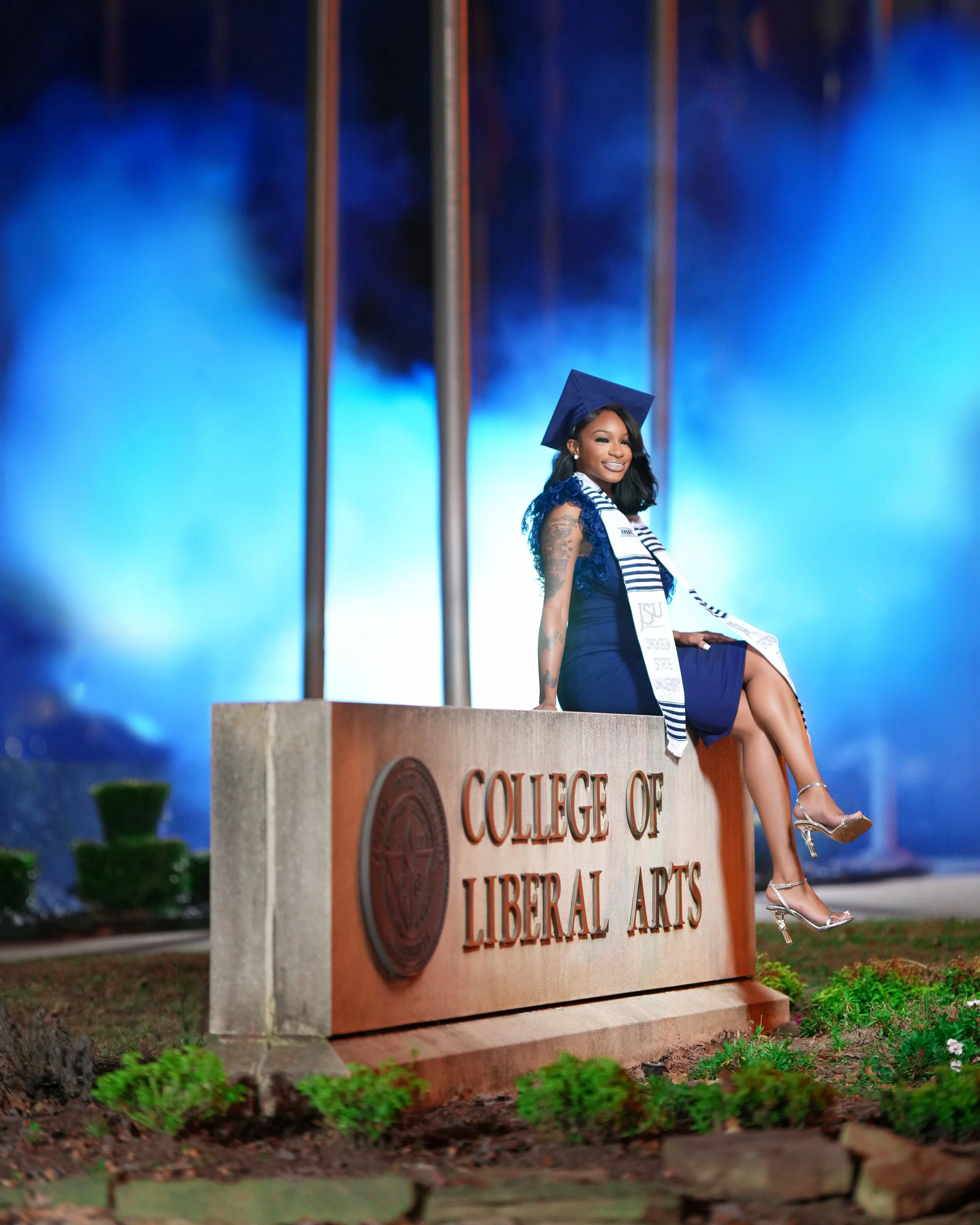 A woman in a blue graduation cap and gown sitting on a sign that reads 'College of Liberal Arts' at a graduation ceremony.