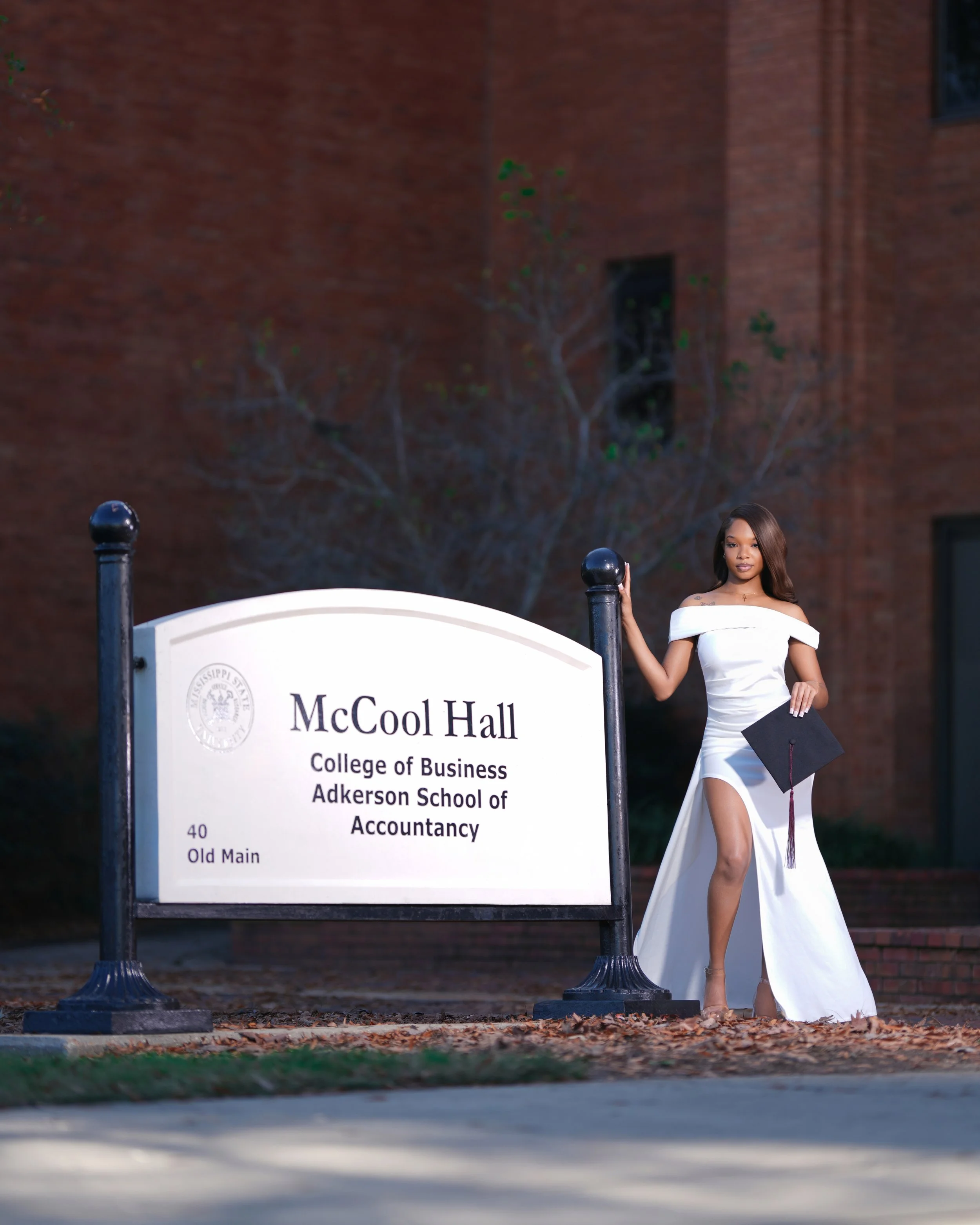 A young woman in a white dress standing next to a sign for McCool Hall at Adkerson School of Accountancy, holding a diploma and a graduation cap.