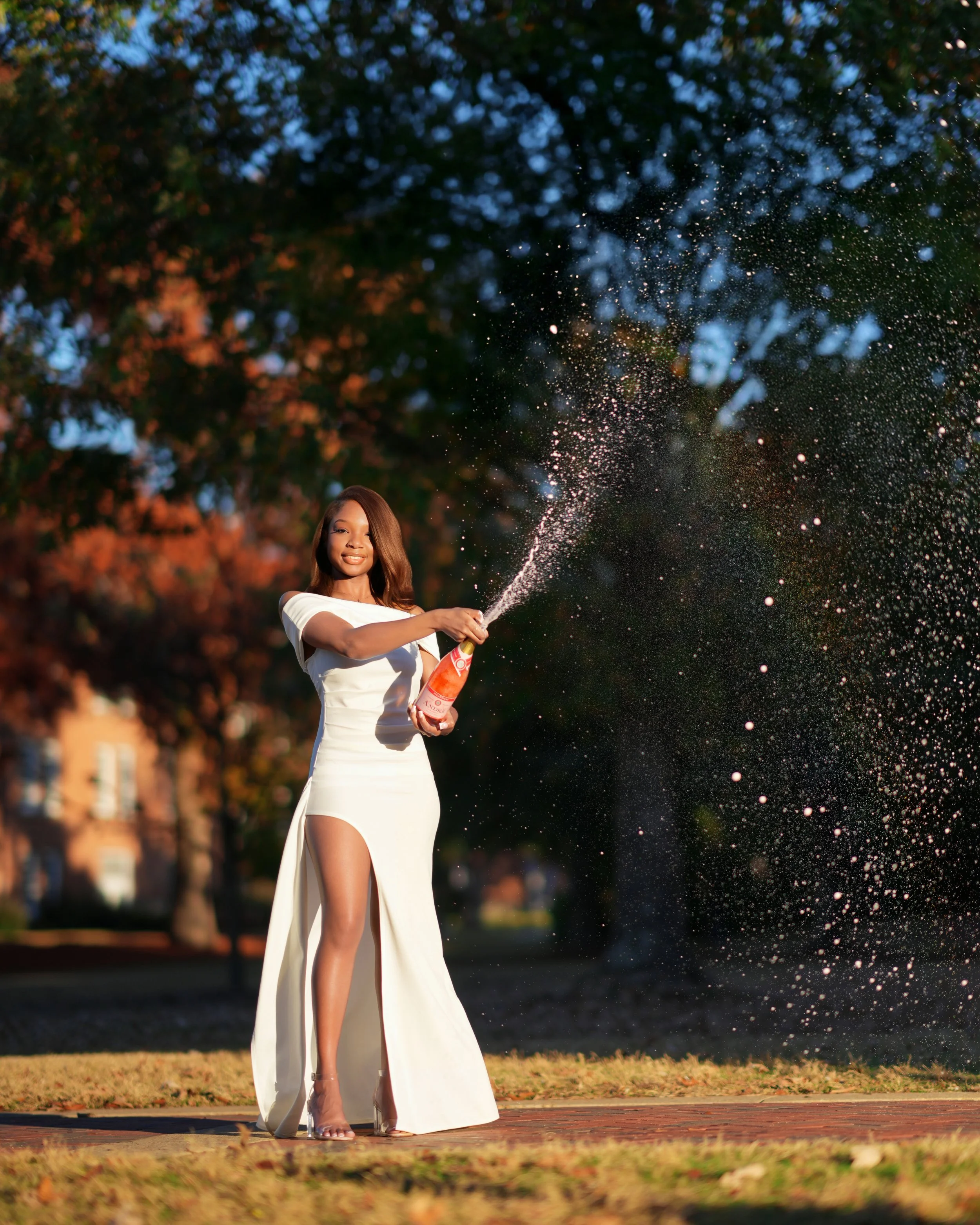 Woman in a white dress opens a bottle of champagne outdoors, causing it to spray, with autumn trees in the background.