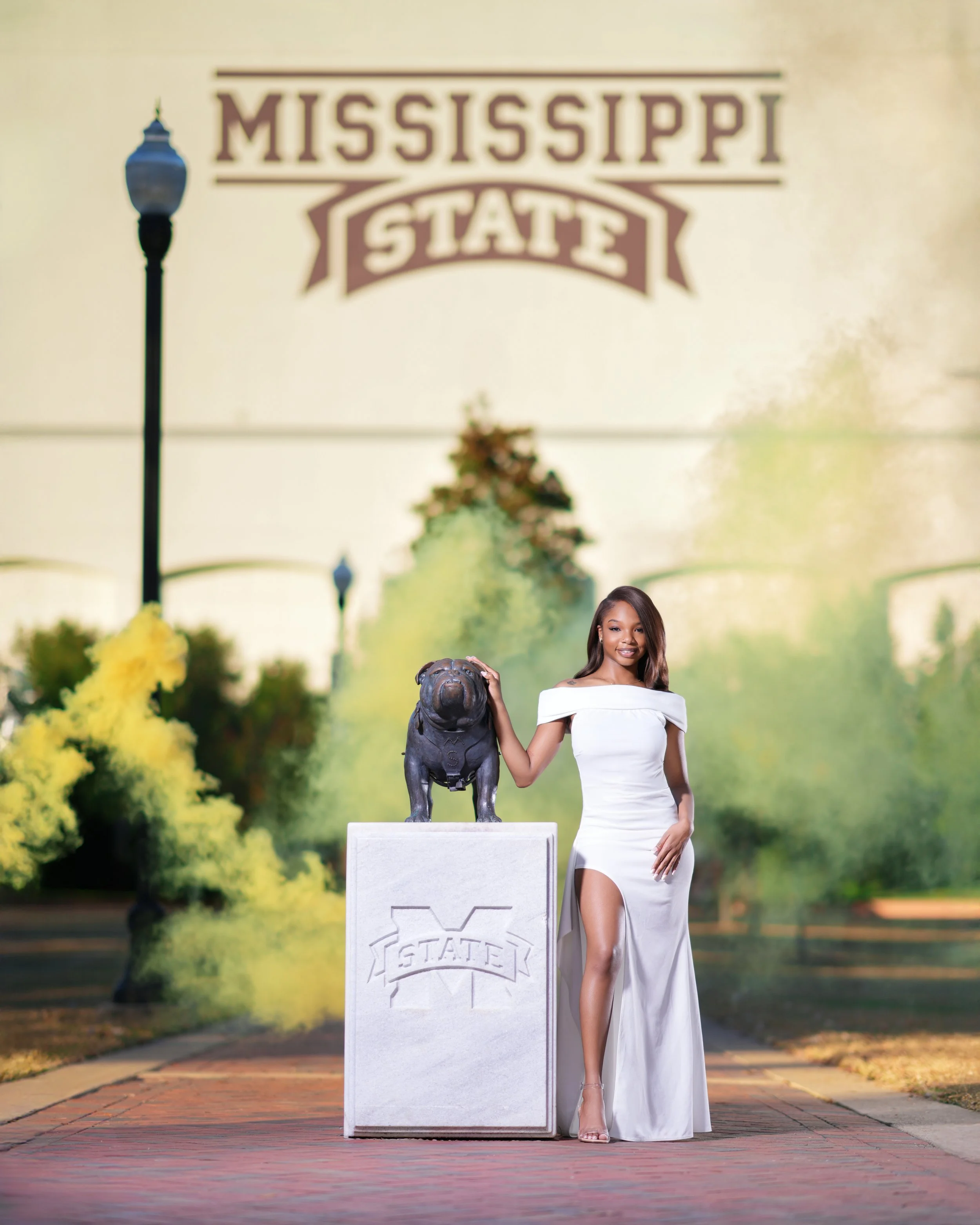 Young woman in a white dress standing next to a statue of a bulldog on a pedestal, with a Mississippi State sign in the background.