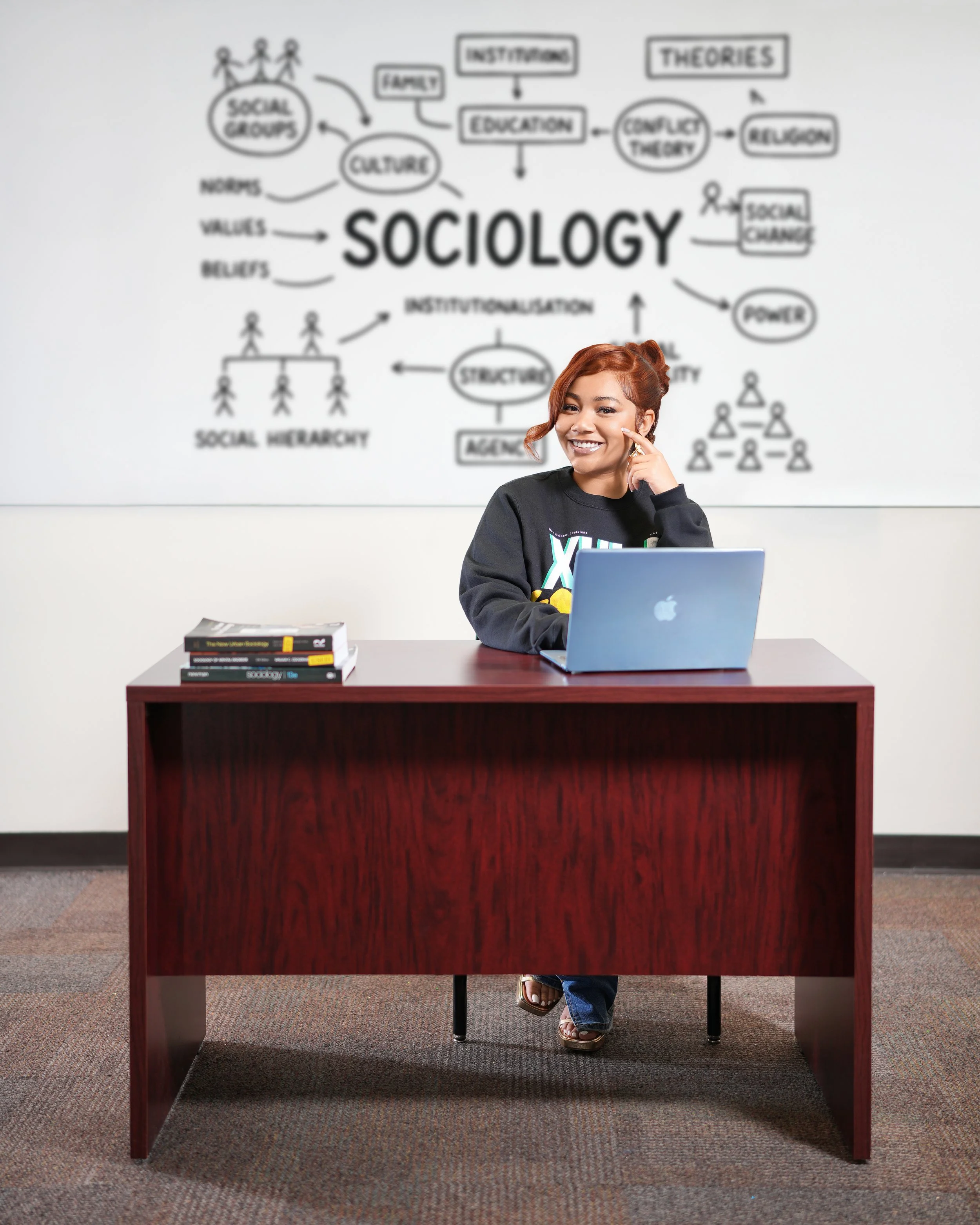 Woman sitting at a desk with a laptop, smiling and touching her face, in front of a whiteboard with a diagram about sociology concepts.
