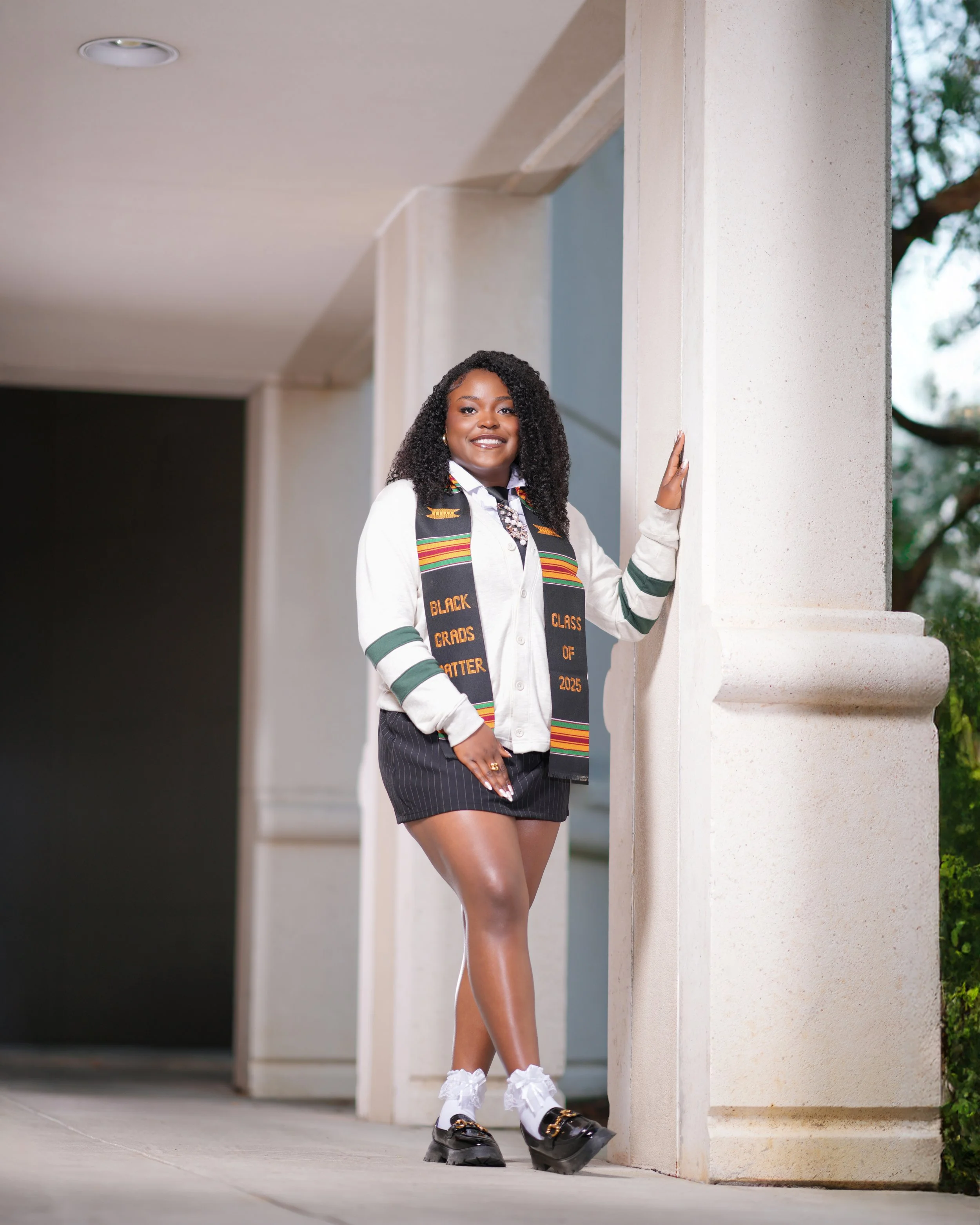 A woman standing outdoors near a building wall, dressed in graduation attire with a stole that reads 'Black Graduates Class of 2025' and a white blouse, black shorts, and loafers, smiling at the camera.