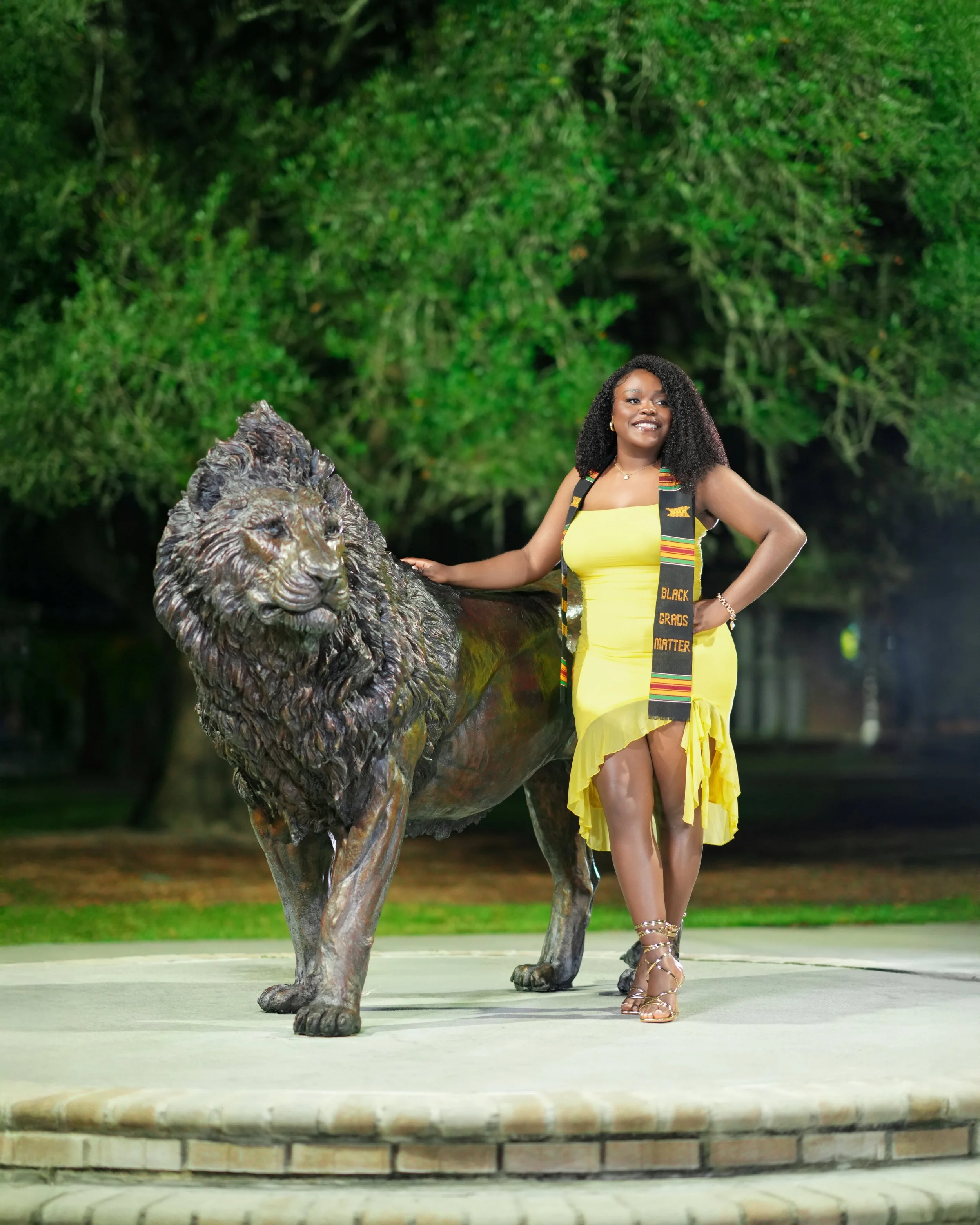A woman in a yellow dress with a Black Lives Matter scarf standing next to a lion statue in a park at night.