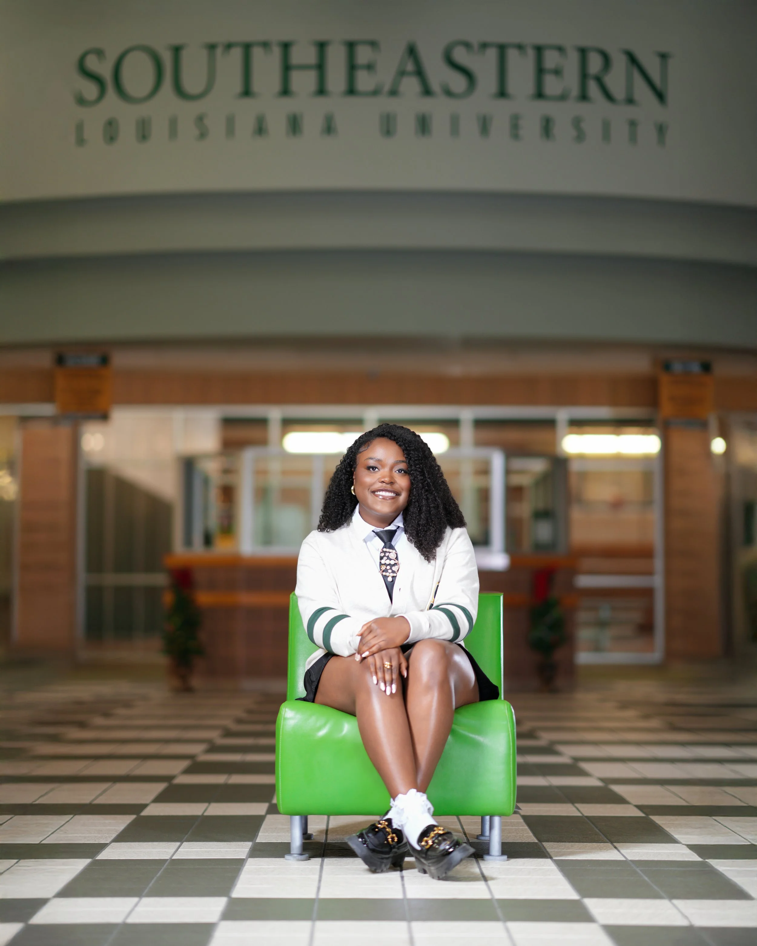 A young woman sitting on a bright green chair inside Southeastern Louisiana University, smiling at the camera, with the university sign visible above in the background.