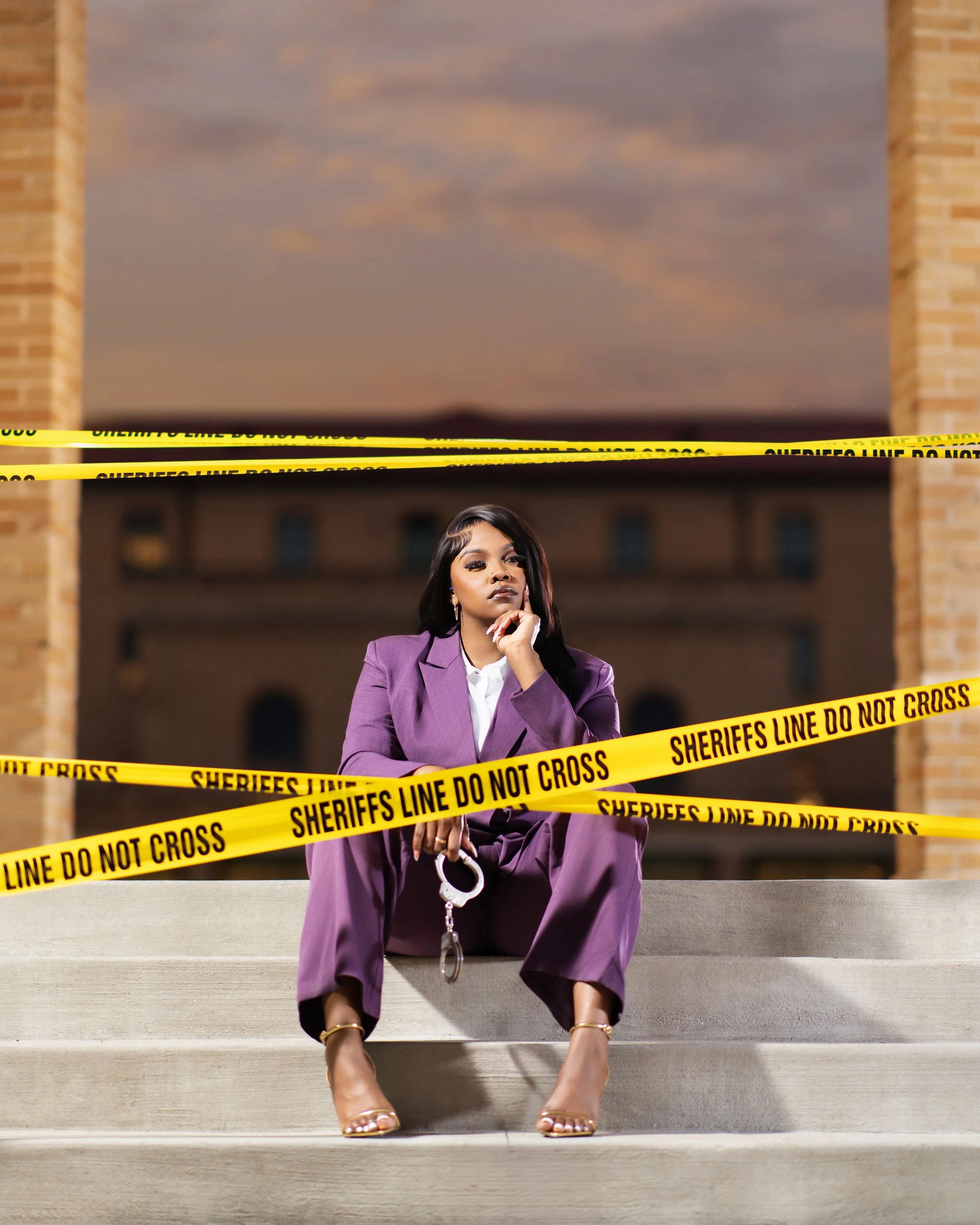 A woman in a purple suit sitting on steps behind yellow police tape reading 'Sheriffs line do not cross,' with a building and evening sky in the background.
