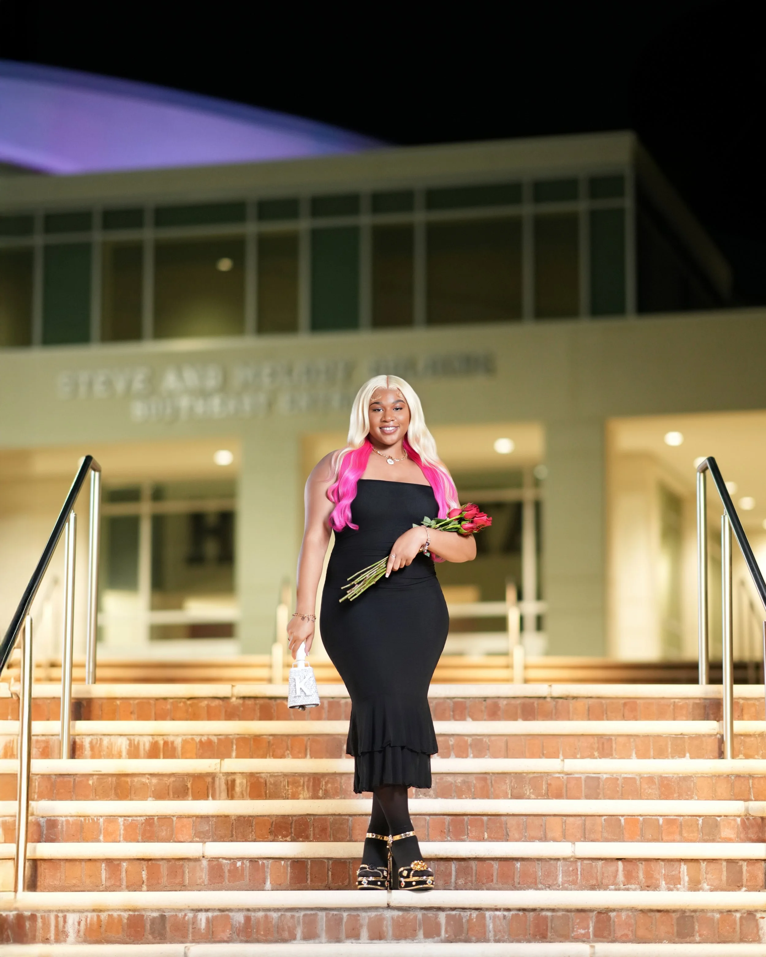 A woman in a black dress holding a bouquet of red roses and a silver handbag stands on steps outside a building at night.