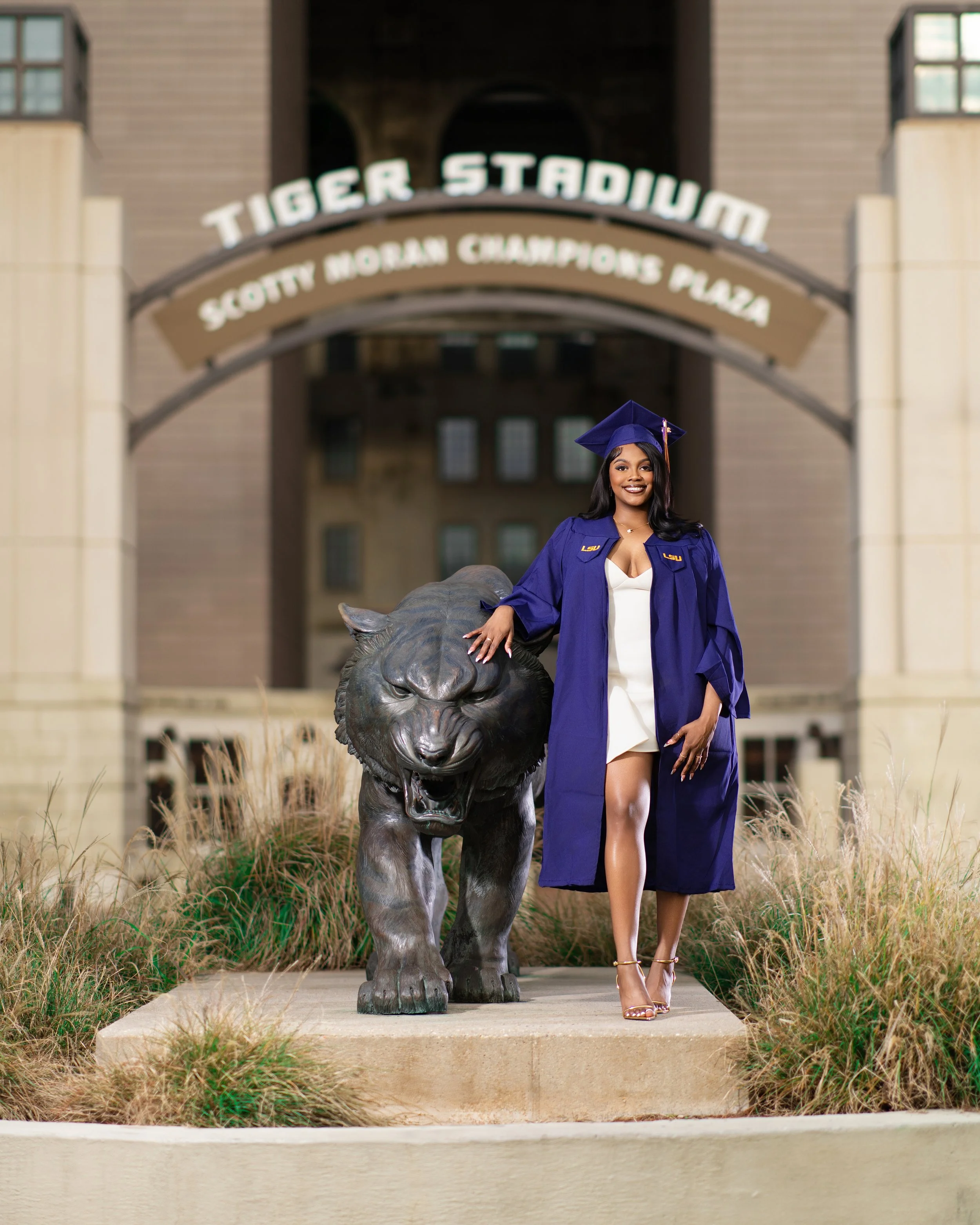 A woman in graduation attire, wearing a blue gown and cap, standing next to a large lion statue in front of Tiger Stadium at Louisiana State University.