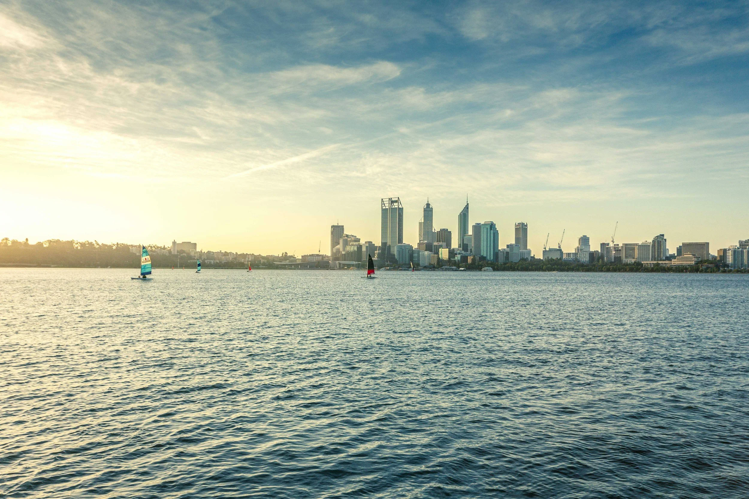 Skyline of Perth city by the water with sailboats and high-rise buildings under a partly cloudy sky.