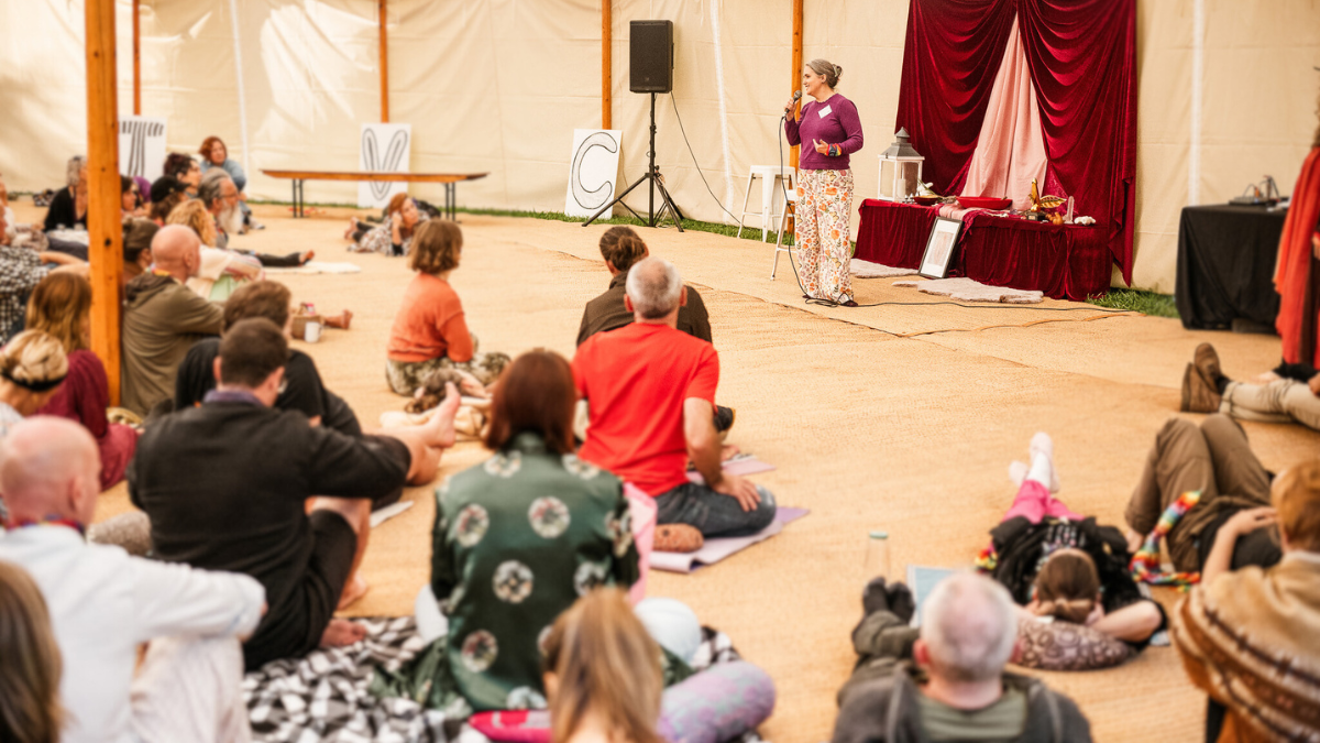 Facilitator leading a communal embodiment workshop inside a tented event space with seated participants.