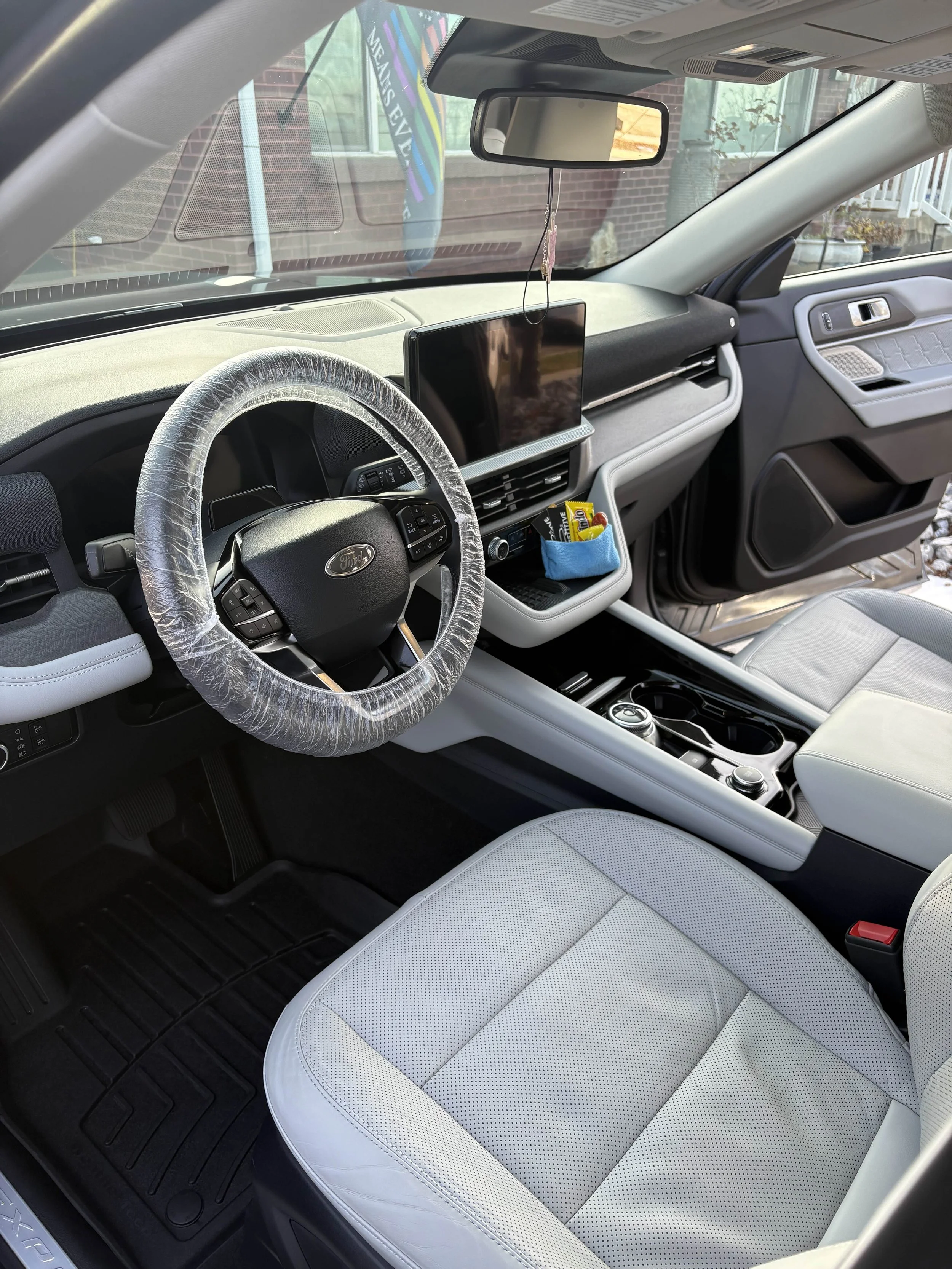 Interior of a car with a plastic wrapped steering wheel, a central touchscreen display, a blue pouch with snacks, and a gear shift in the center console.