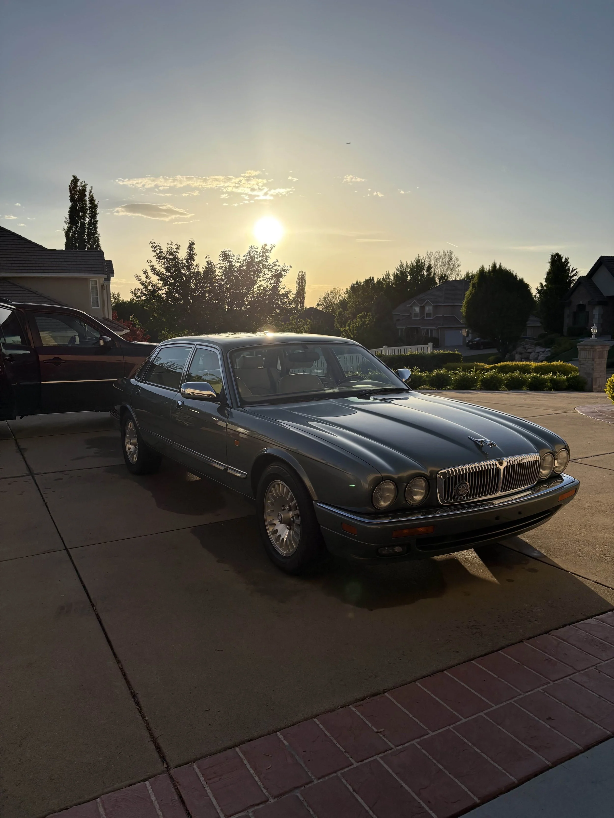 A classic gray Jaguar XJ-S coupe parked in a driveway during sunset with a suburban neighborhood and trees in the background.