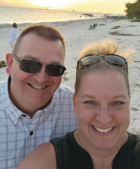 A smiling man and woman taking a selfie on a beach at sunset, with beachgoers and boats in the background.