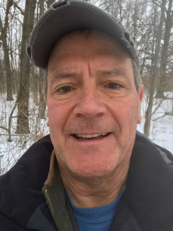 A man outdoors in a snowy forest, wearing a gray baseball cap and a black jacket, smiling at the camera.