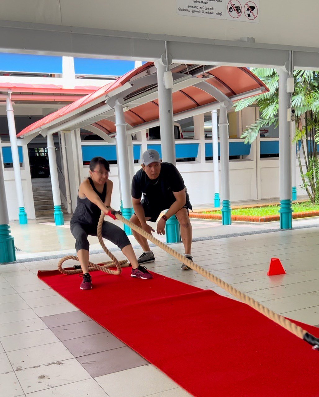 Two people engaged in a tug-of-war on a red carpet inside a covered area with a white tiled floor and turquoise pillars.