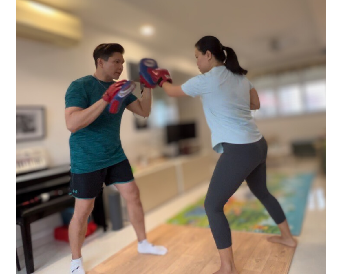 Two individuals practicing boxing in a living room, one wearing focus mitts and the other punching, both dressed in casual workout attire.
