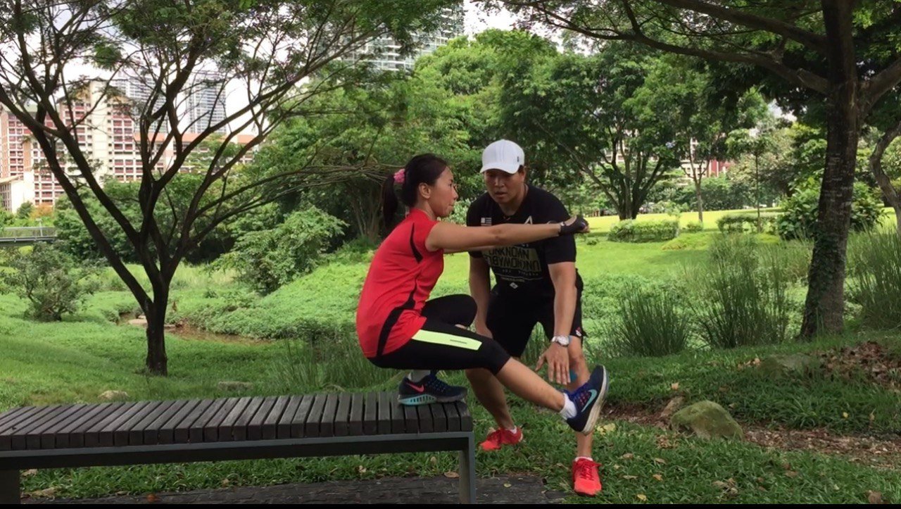 Two people exercising on a bench in a park surrounded by trees and greenery.