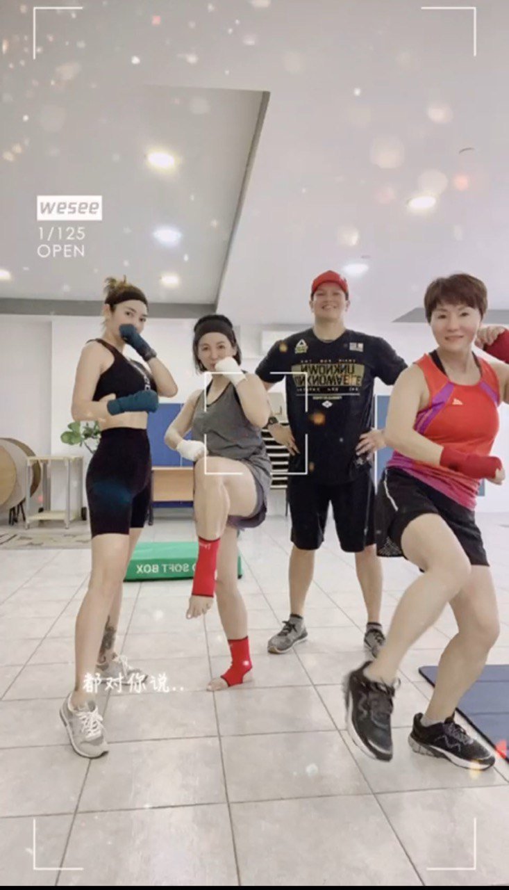 Group of four people posing in a gym setting, wearing athletic clothing and showing martial arts or boxing stances. All are smiling and appear energetic. The background shows gym equipment, including soft boxes and exercise mats.